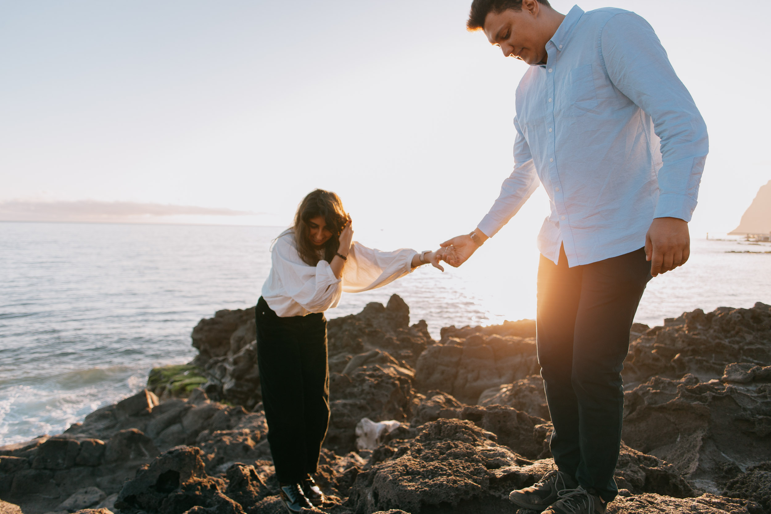 Man helping woman across volcanic rocks at sunset at Praia Formosa, holding hands, Funchal cliffs with houses behind, golden backlight, Madeira
