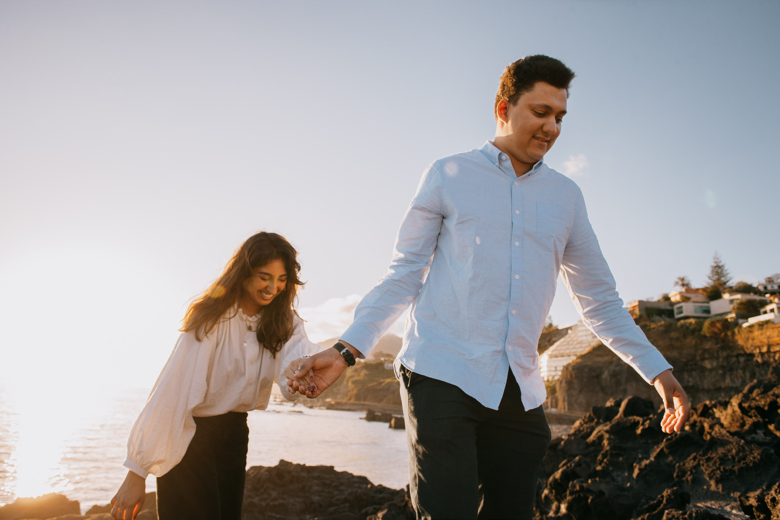 Man guiding woman by hand across volcanic rocks, she smiling down at her footing, Funchal cliffs with houses behind, golden light, Praia Formosa, Madeira