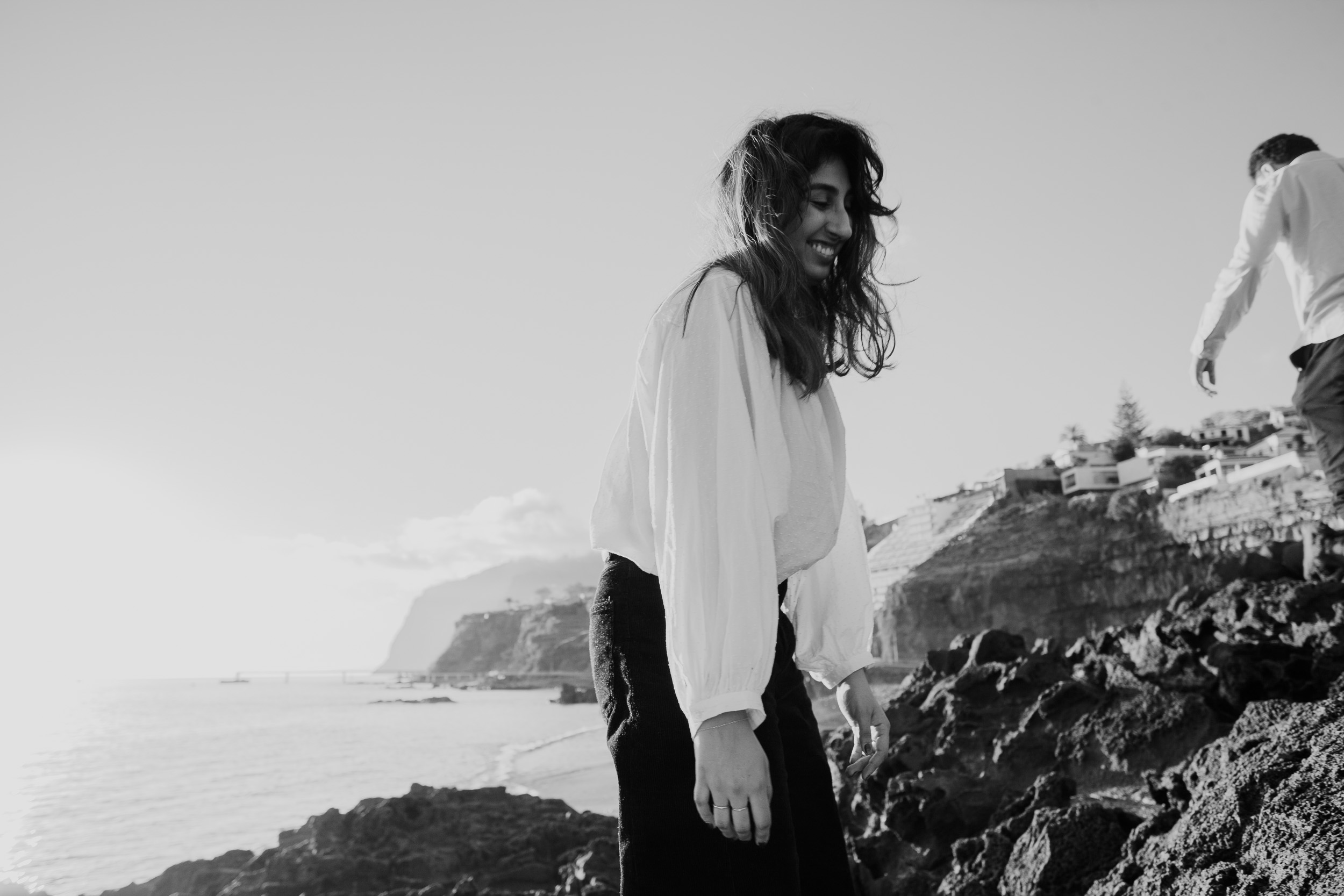 Black and white portrait of woman smiling with hair blowing in wind at Praia Formosa, Funchal cliffs and Atlantic behind, Madeira