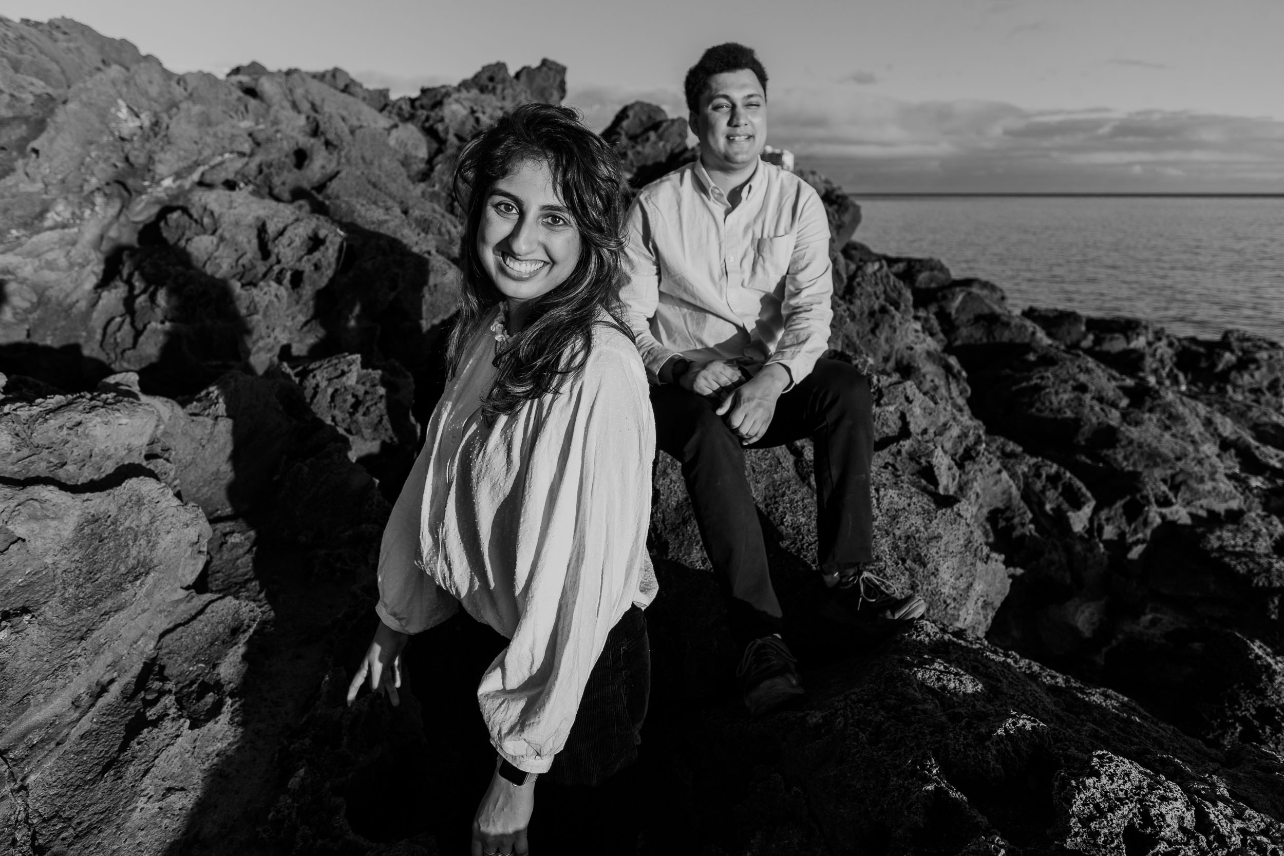 Black and white portrait of woman seated on volcanic rocks looking back at camera smiling, man seated above behind her, Atlantic behind, Praia Formosa, Madeira