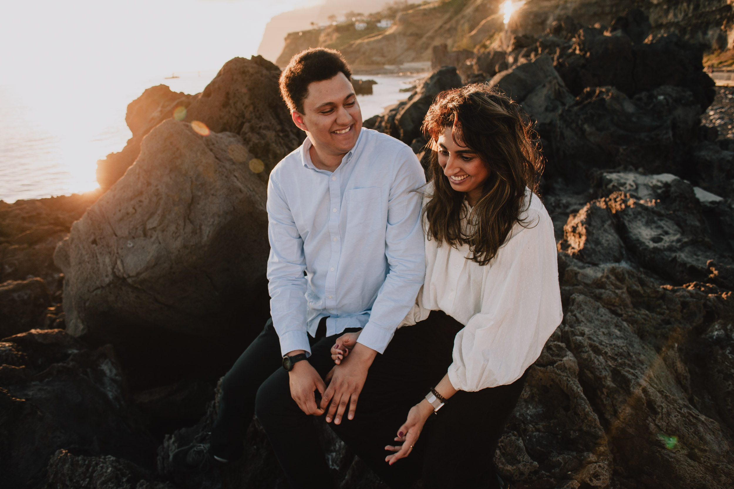 Couple seated on volcanic rocks sharing a laugh, man smiling at her, she smiling down, golden sunset light, Funchal cliffs behind, Praia Formosa, Madeira