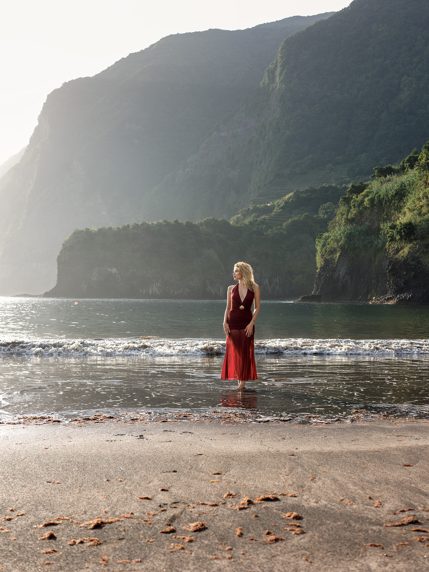 Woman in red halter dress standing in shallow water at Seixal Beach, volcanic cliffs and morning haze behind, Madeira
