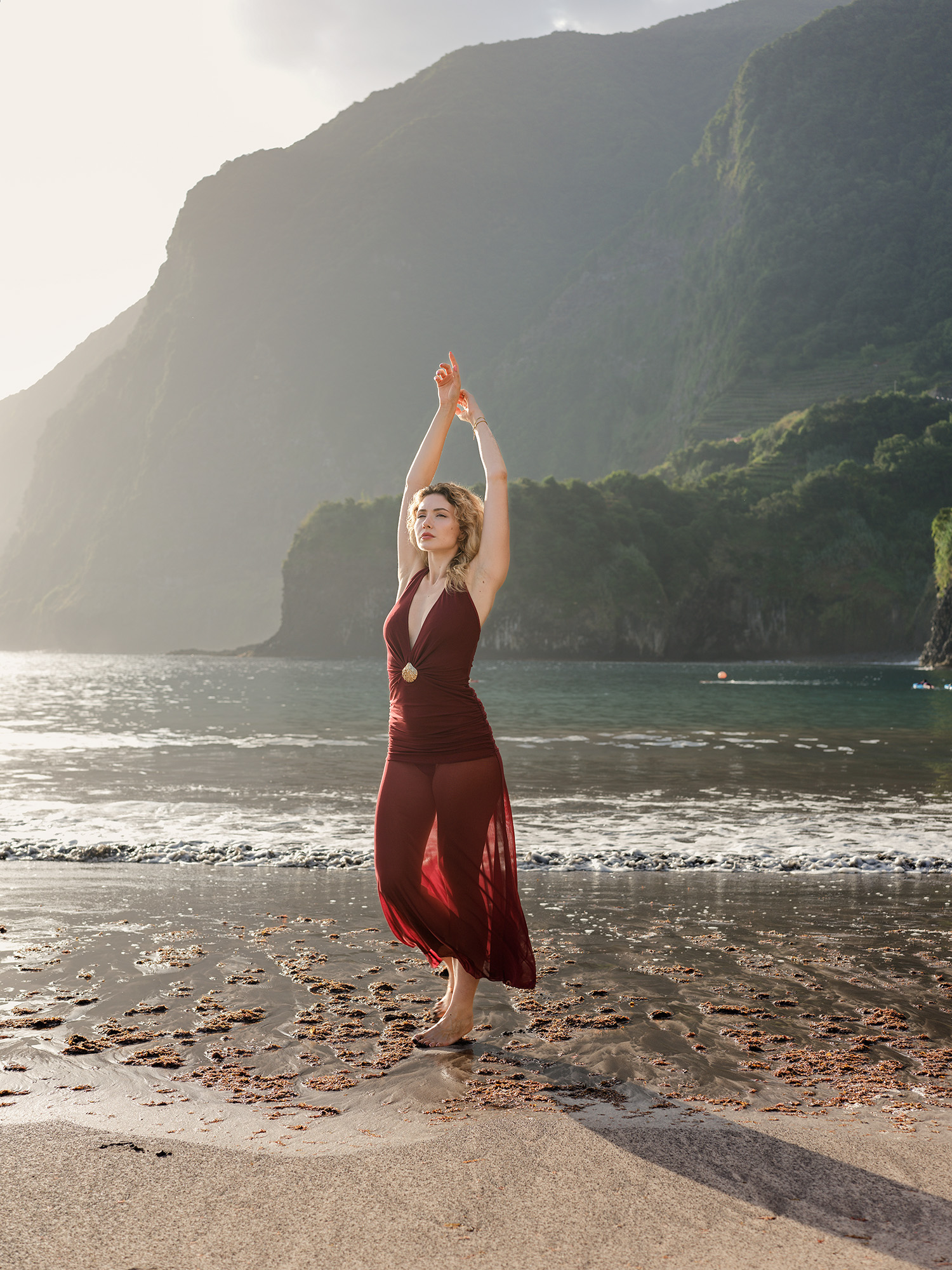 Woman in red dress with arms raised on Seixal Beach, morning cliffs and Atlantic behind, Madeira