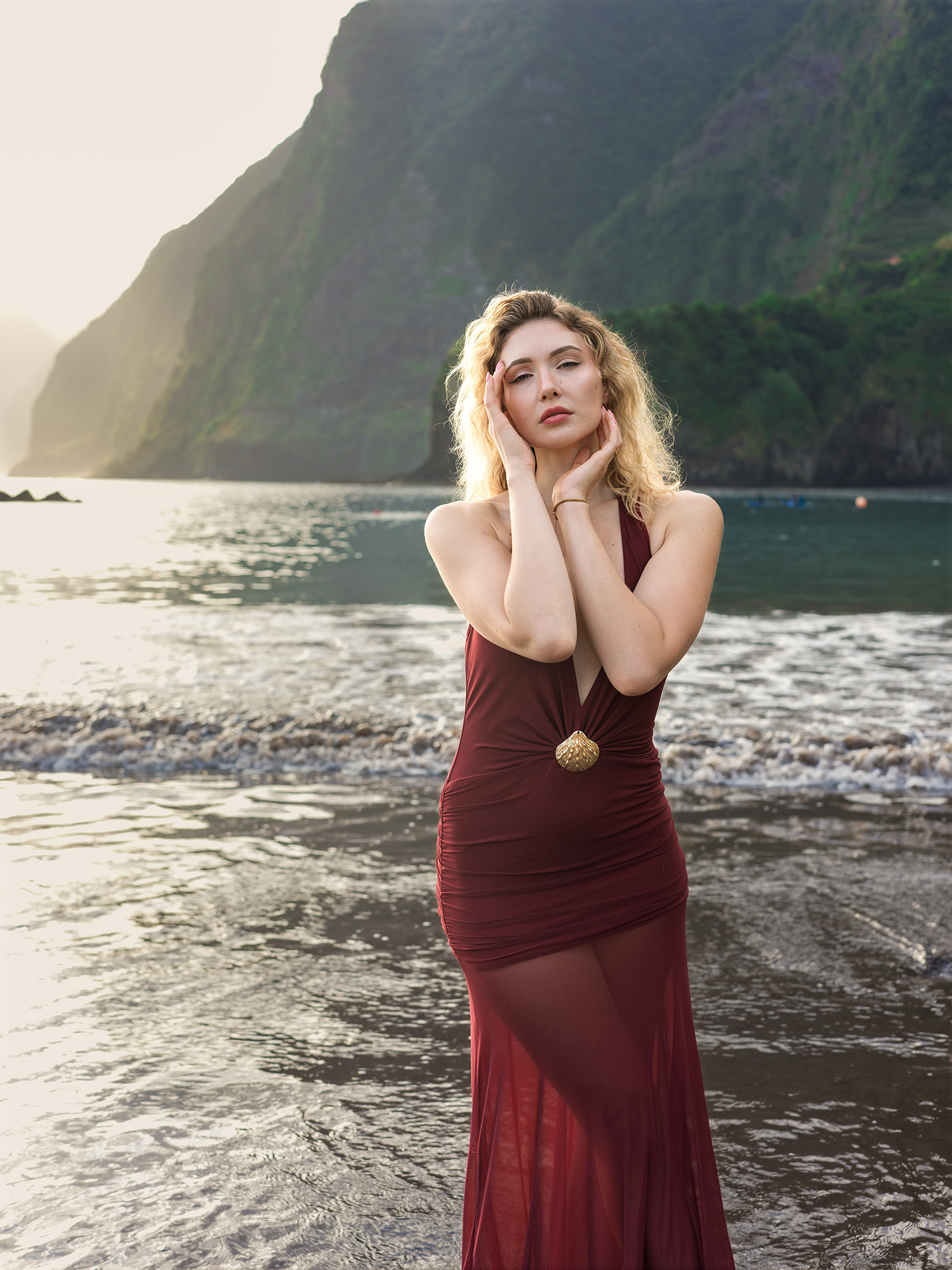 Full-length portrait in the red halter dress, both hands raised to her face, the green headland and the lit water behind, Seixal Beach, Madeira