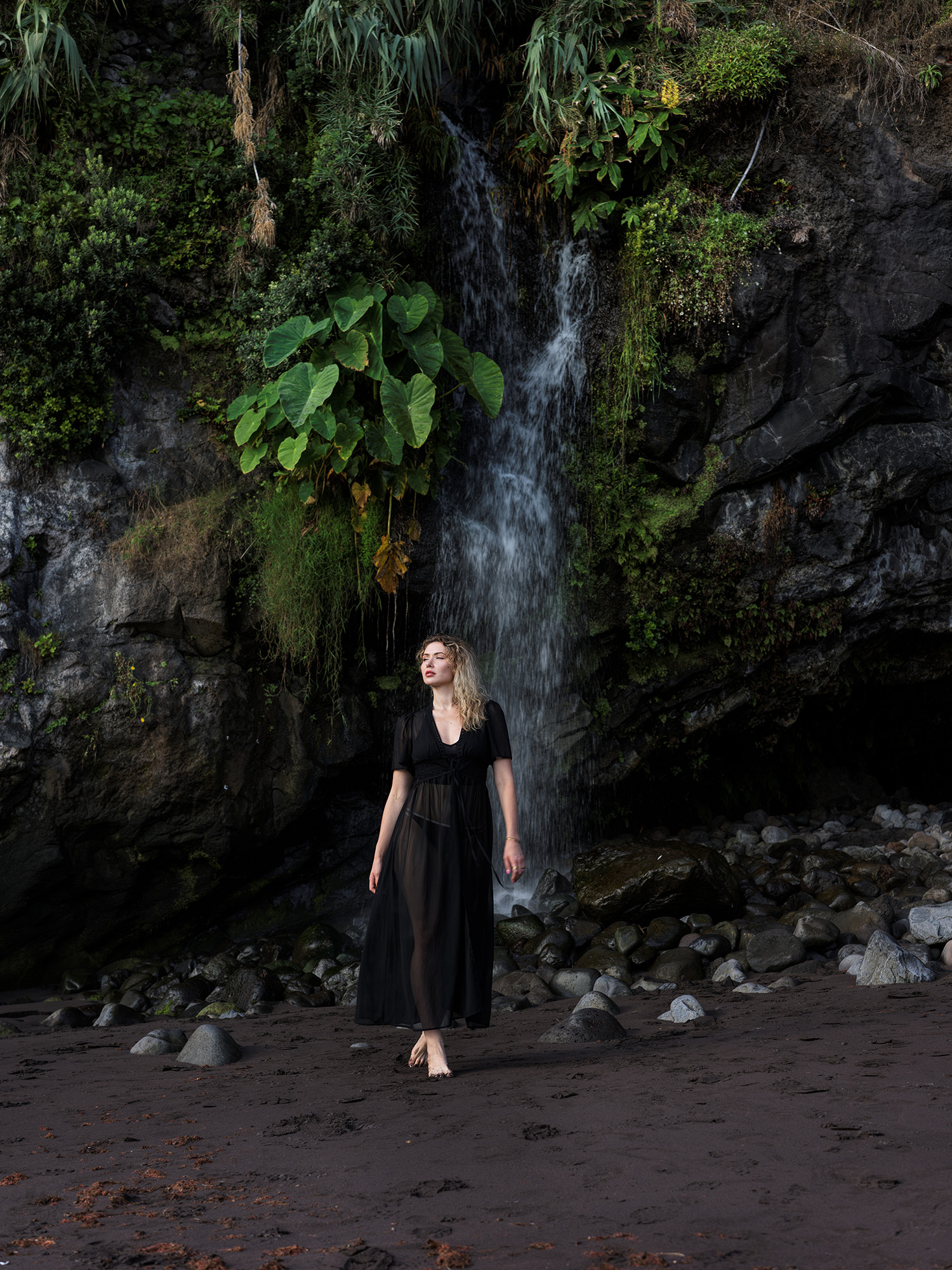 Woman in black dress standing before a waterfall on dark cliff face, lush vegetation above, Seixal Beach, Madeira