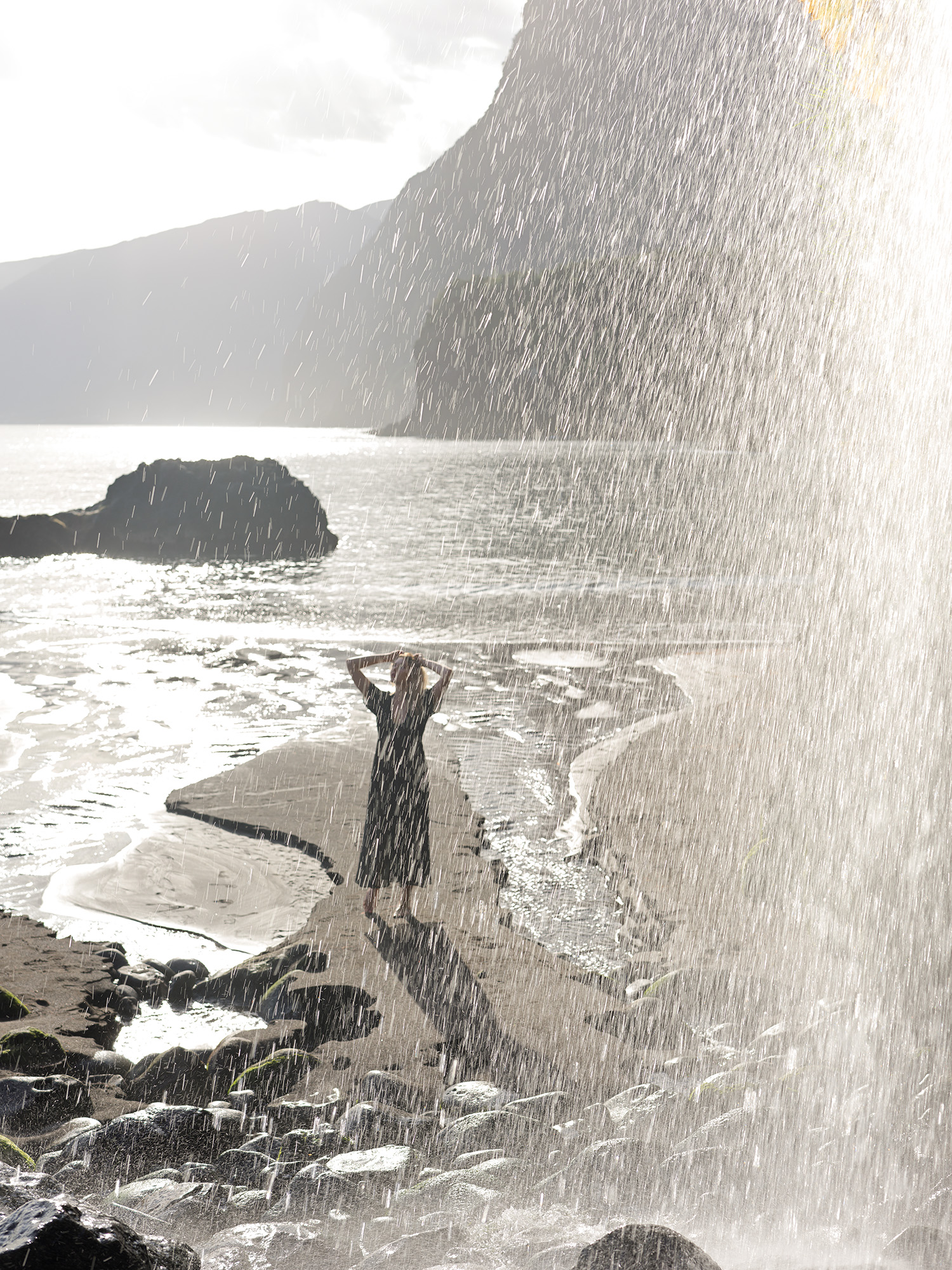 Woman standing under waterfall spray at Seixal Beach, black dress, cliffs and Atlantic behind, backlit, Madeira