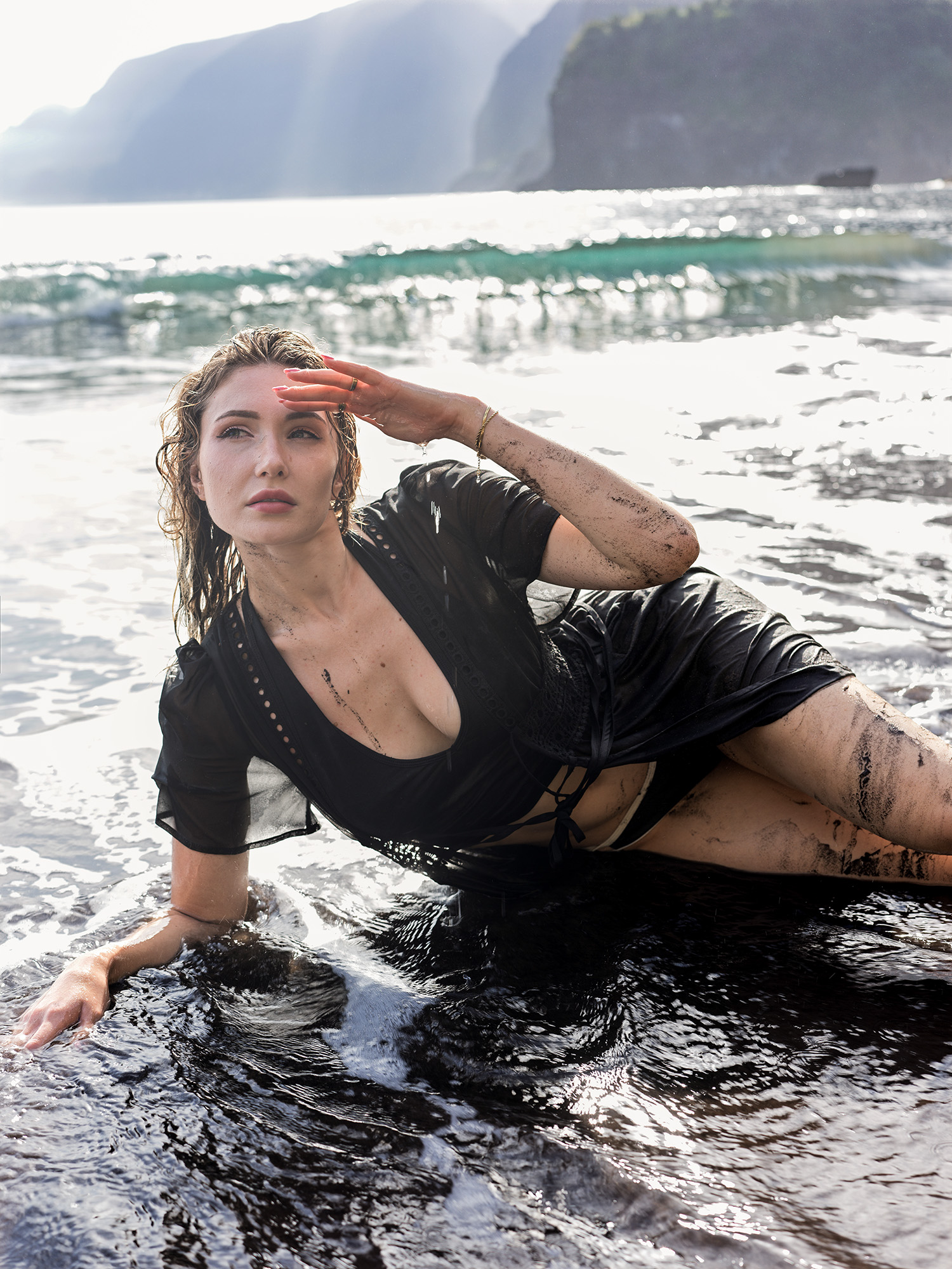 Woman in wet black dress lying in shallow water on black volcanic sand at Seixal Beach, cliffs behind, Madeira