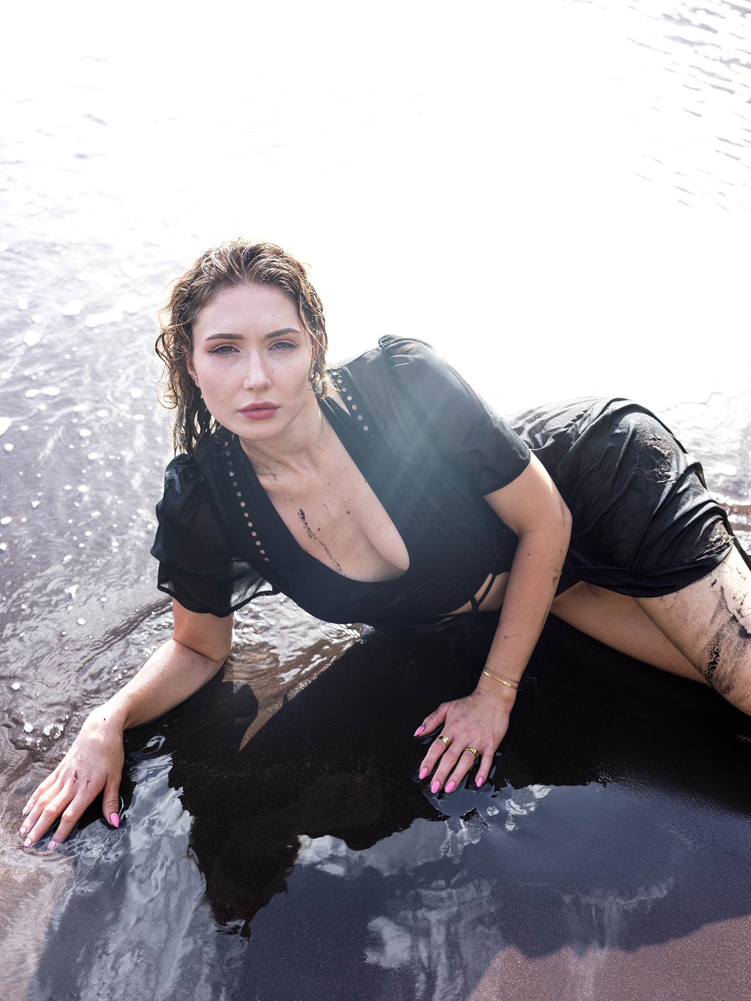 Woman in wet black dress lying on black volcanic sand at Seixal Beach, looking directly at camera, Madeira