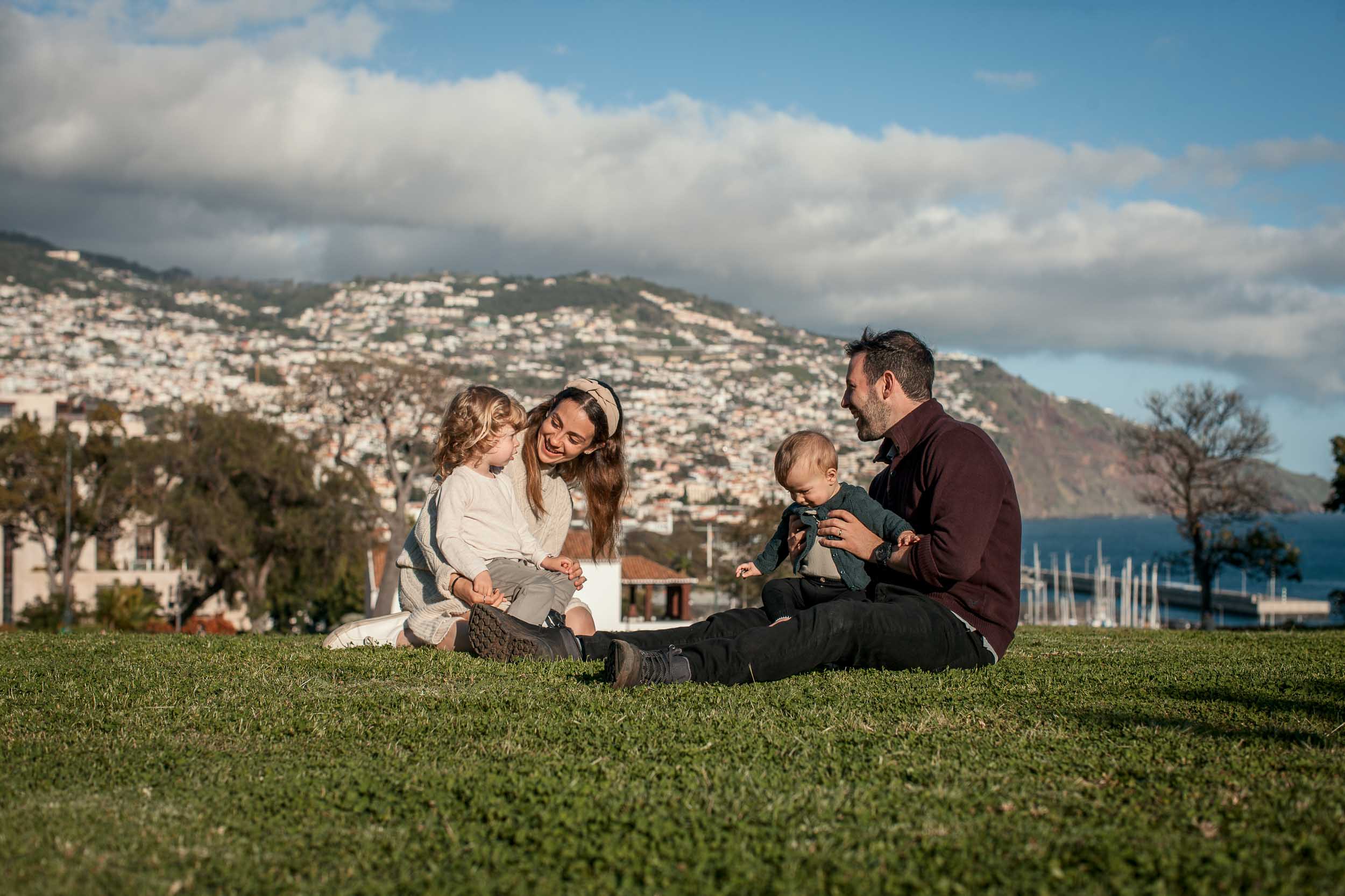 Family of four sitting on the lawn at Santa Catarina Park, parents facing both children, Funchal cityscape and harbor behind, sunny, Madeira