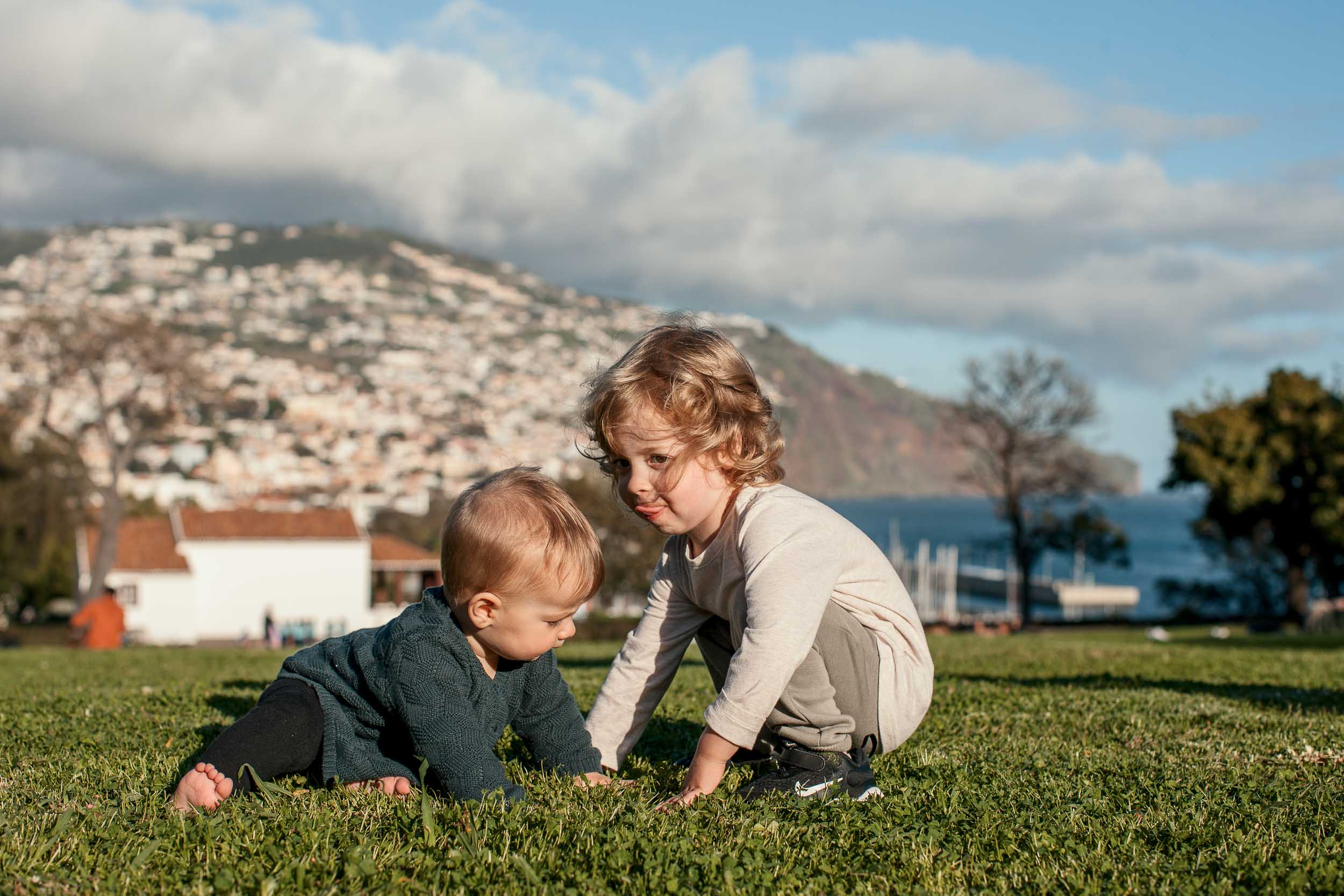 Baby and curly-haired toddler playing face to face on the grass at Santa Catarina Park, Funchal hillside behind, Madeira
