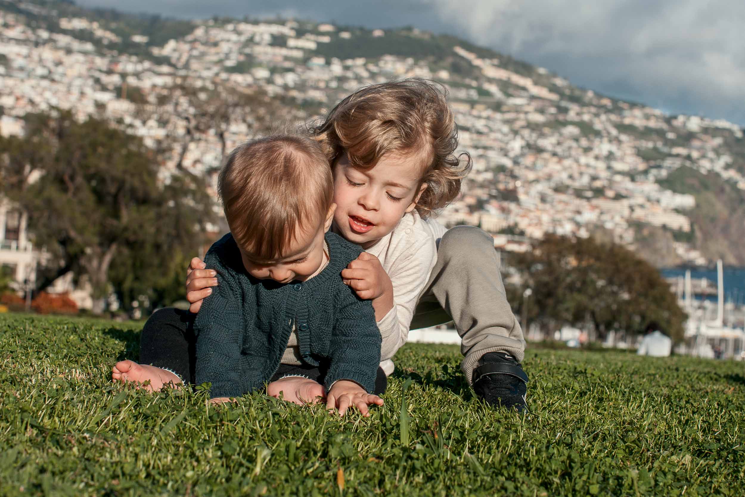 Toddler leaning over and hugging baby from behind on the lawn at Santa Catarina Park, Funchal harbor in the distance, Madeira