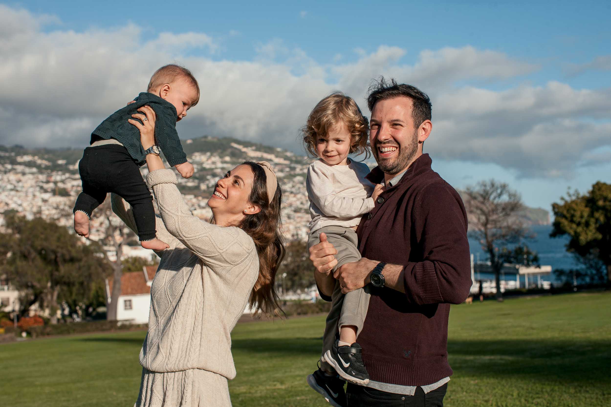 Both parents each lifting a child into the air and laughing, Santa Catarina Park lawn, Funchal cityscape behind, Madeira