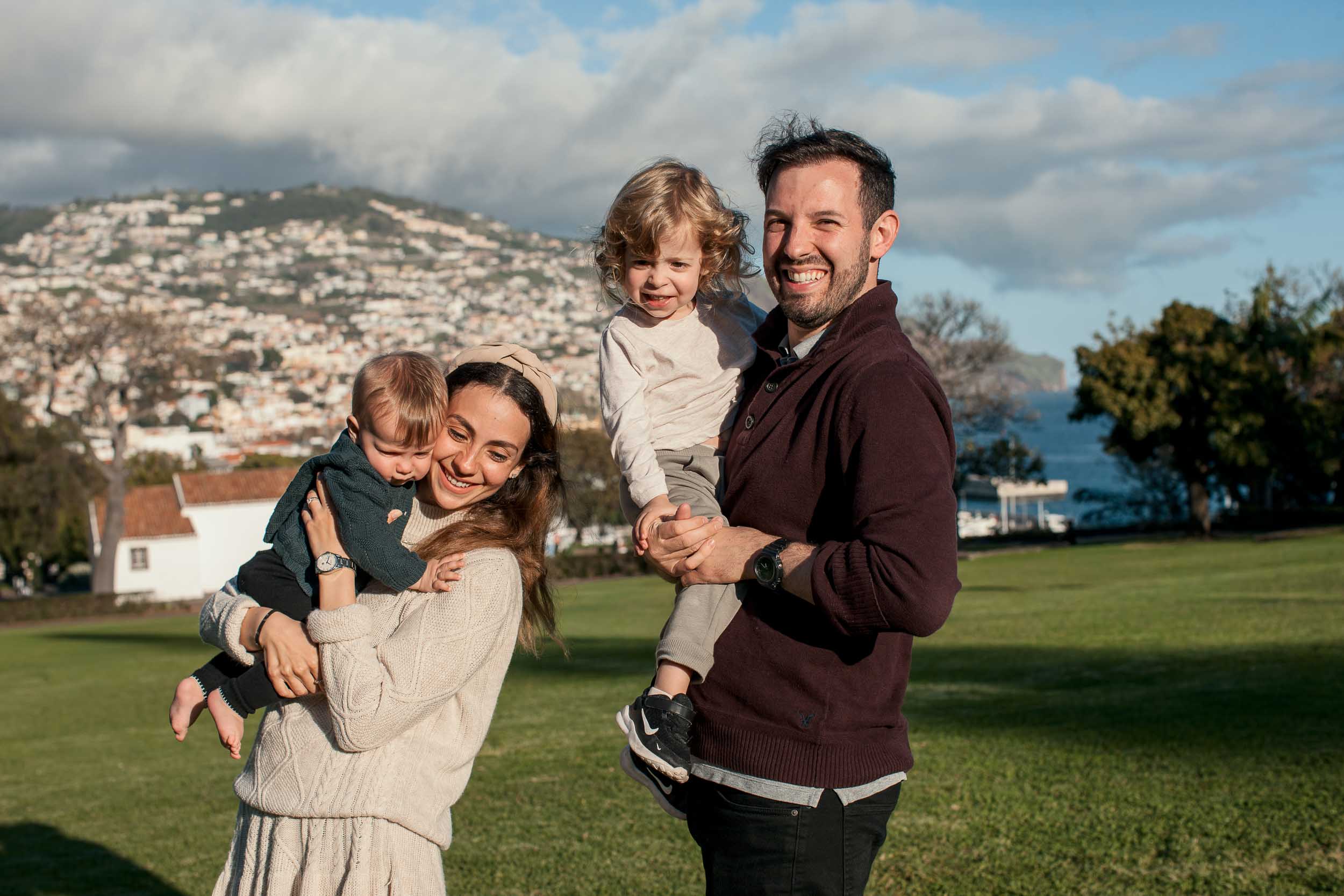 Mother holding baby, father holding toddler on his hip, both parents smiling at camera, Santa Catarina Park, Funchal harbor and Atlantic behind, Madeira