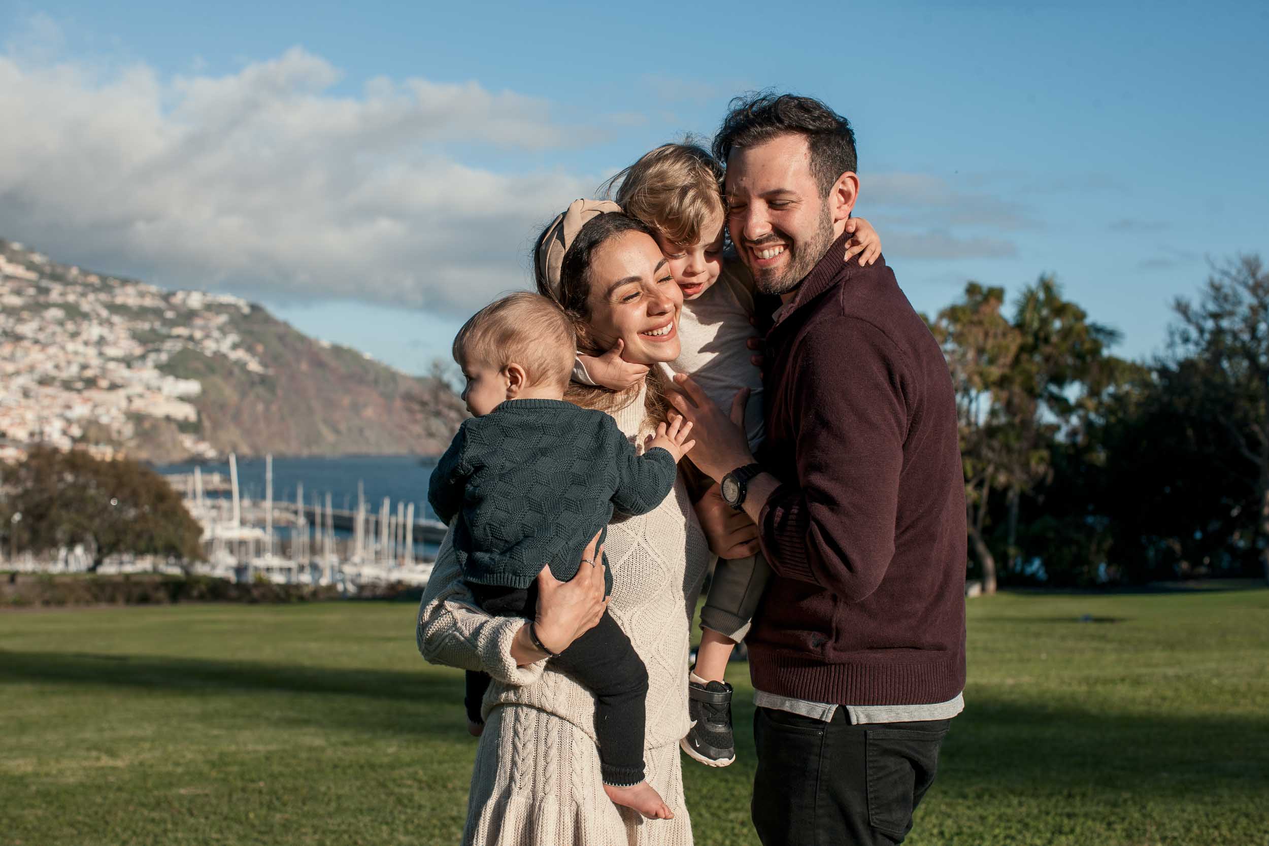 Father leaning in smiling as toddler climbs onto mother's shoulder, mother holding baby, Santa Catarina Park, Funchal marina behind, Madeira