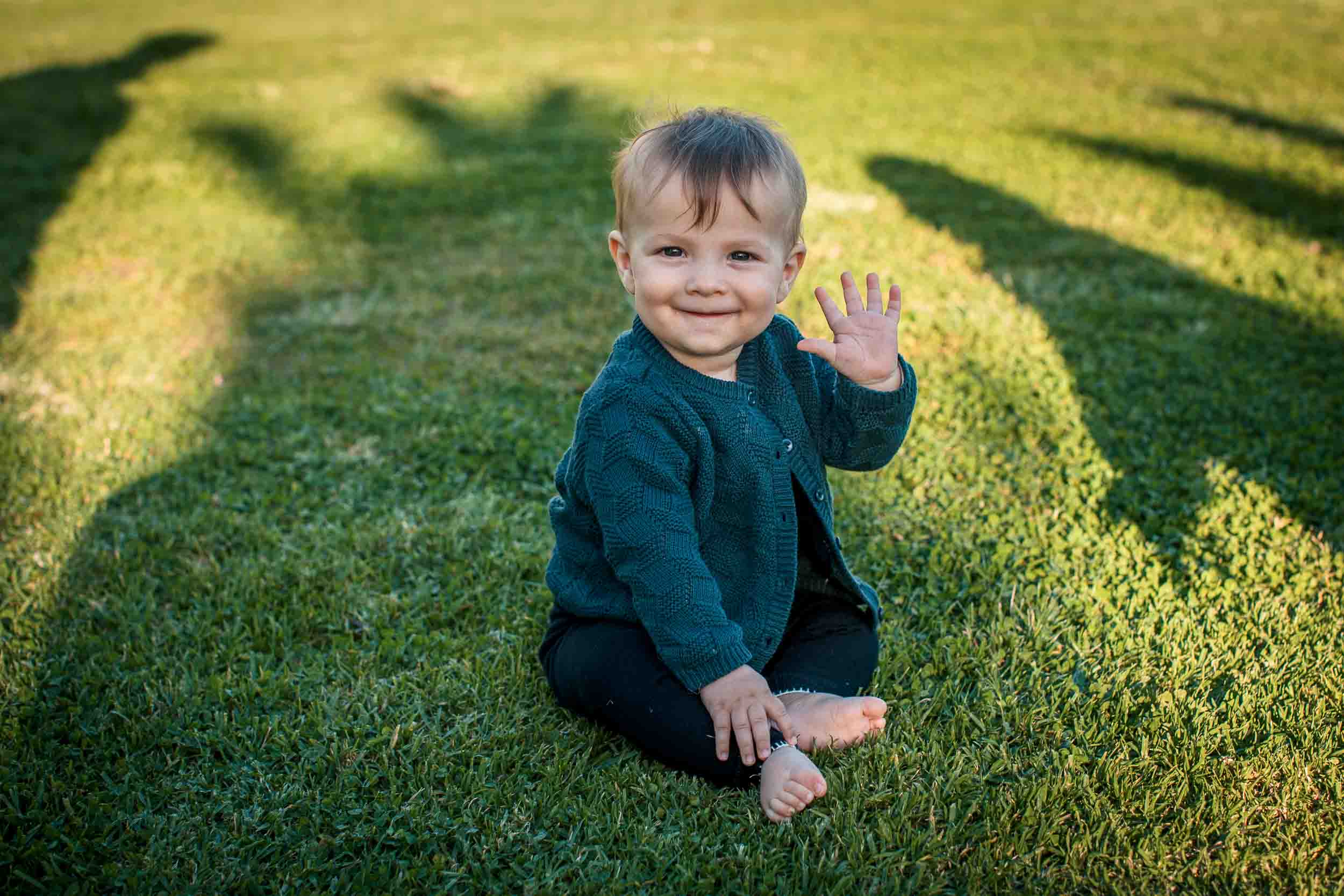 Child sitting on the lawn at Santa Catarina Park, waving at the camera, palm tree shadows across the grass, Funchal, Madeira