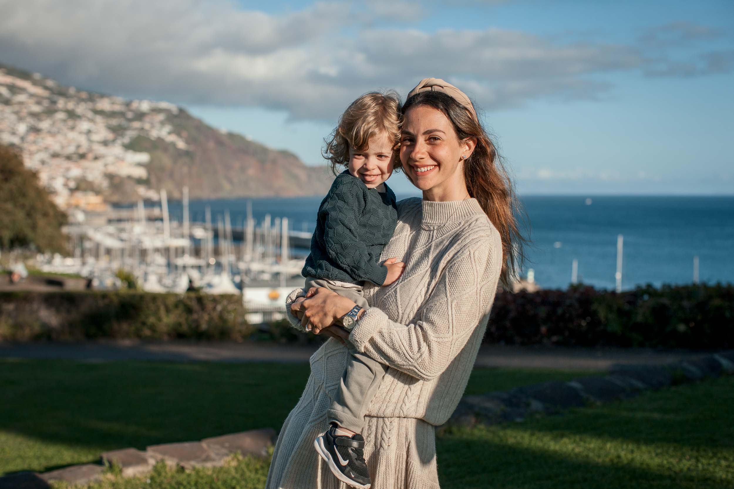 Mother holding up a laughing toddler, both smiling, Santa Catarina Park, Funchal marina and yachts behind, Madeira