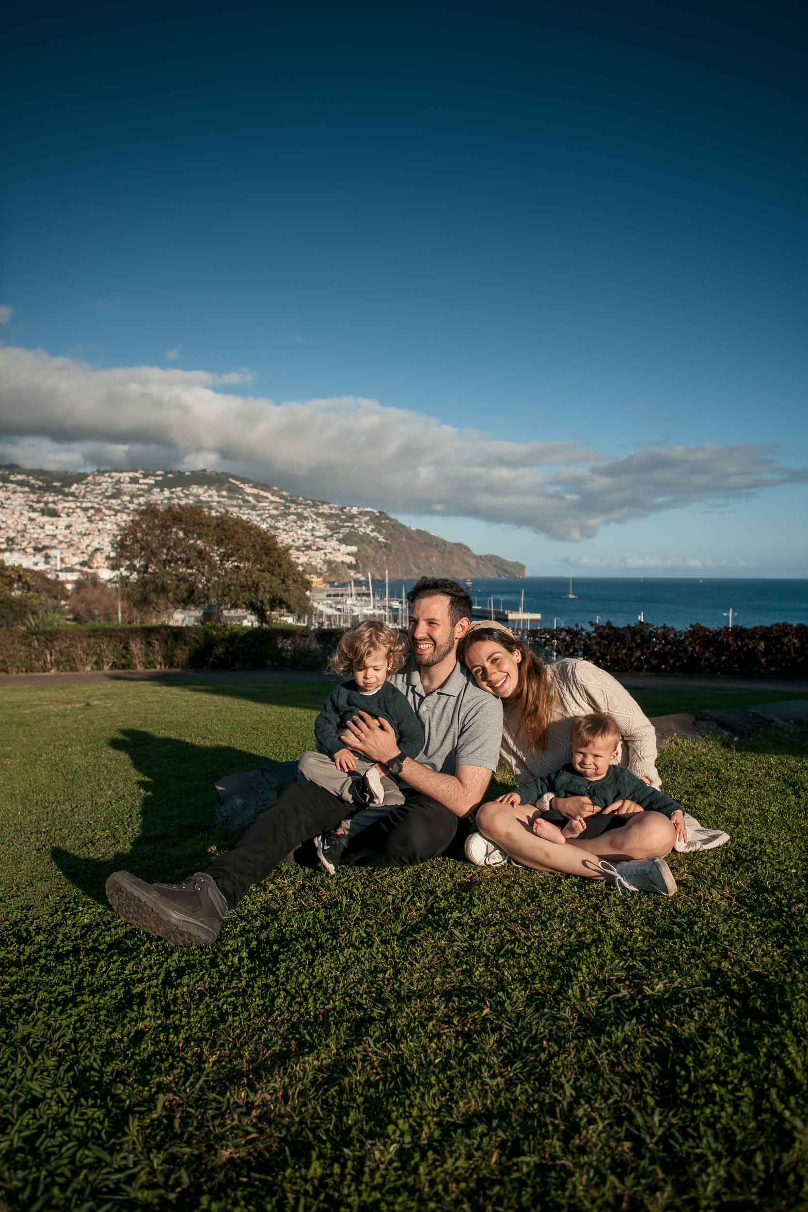 Family of four sitting close together on the lawn at Santa Catarina Park, father laughing, Funchal harbor and marina behind, deep blue sky, Madeira