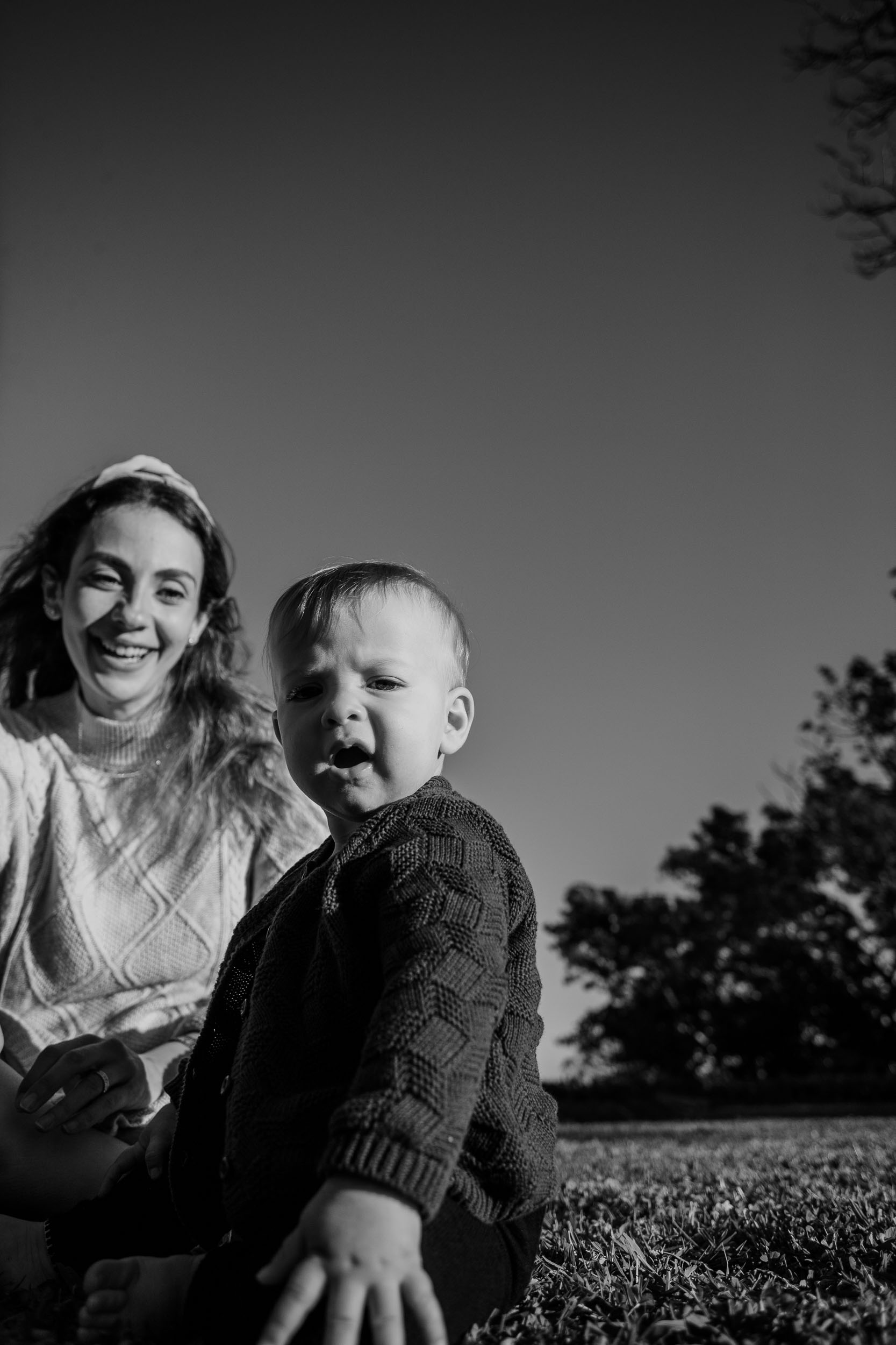 Black and white. mother laughing as baby makes an animated expression very close to camera, park trees and open sky behind, Santa Catarina Park, Madeira