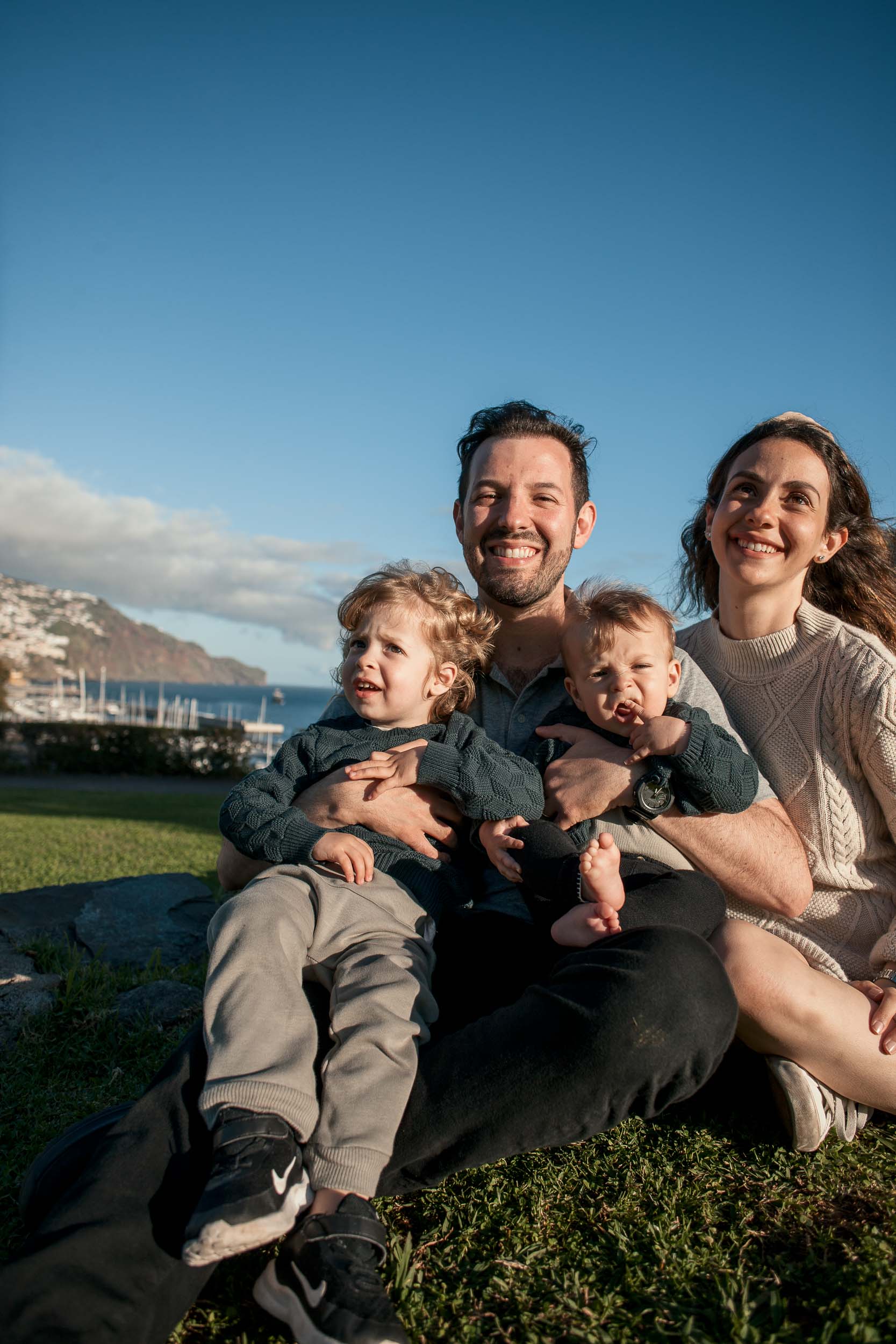 Family of four seated on a low stone wall at Santa Catarina Park, both parents each holding a child, Funchal harbor and marina behind, Madeira