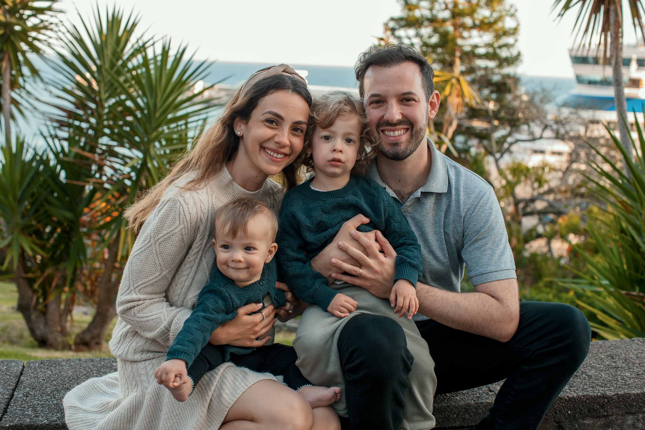 Family of four seated on stone steps at Santa Catarina Park, both children in teal, cruise ship terminal and tall palm trees behind, Funchal, Madeira
