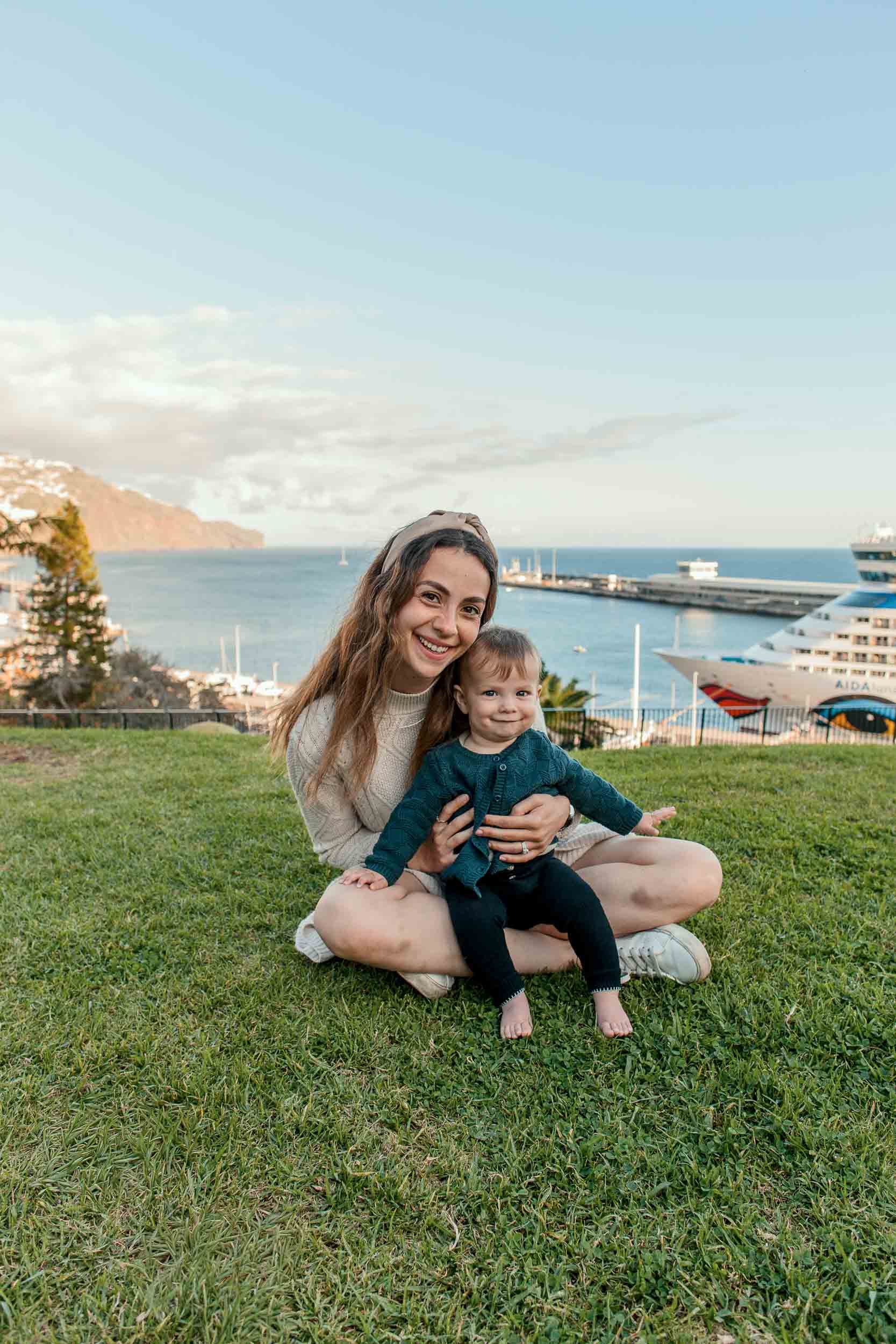 Mother sitting on the lawn at Santa Catarina Park with a child on her lap, Funchal harbor and cruise ship visible behind, Madeira