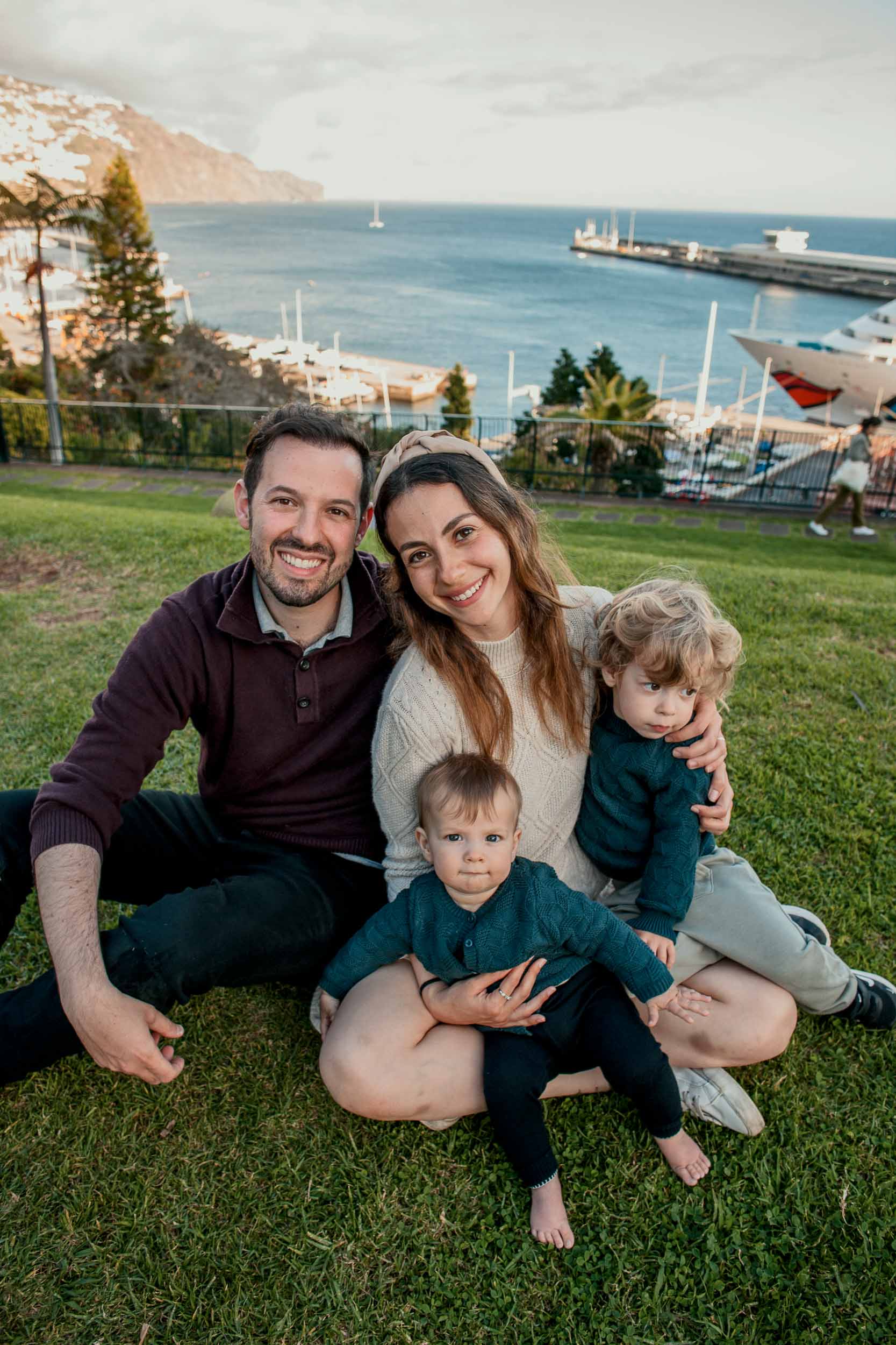 Family of four seated on the lawn hillside at Santa Catarina Park, parents smiling, Funchal marina and Atlantic behind, dramatic cloudy sky, Madeira
