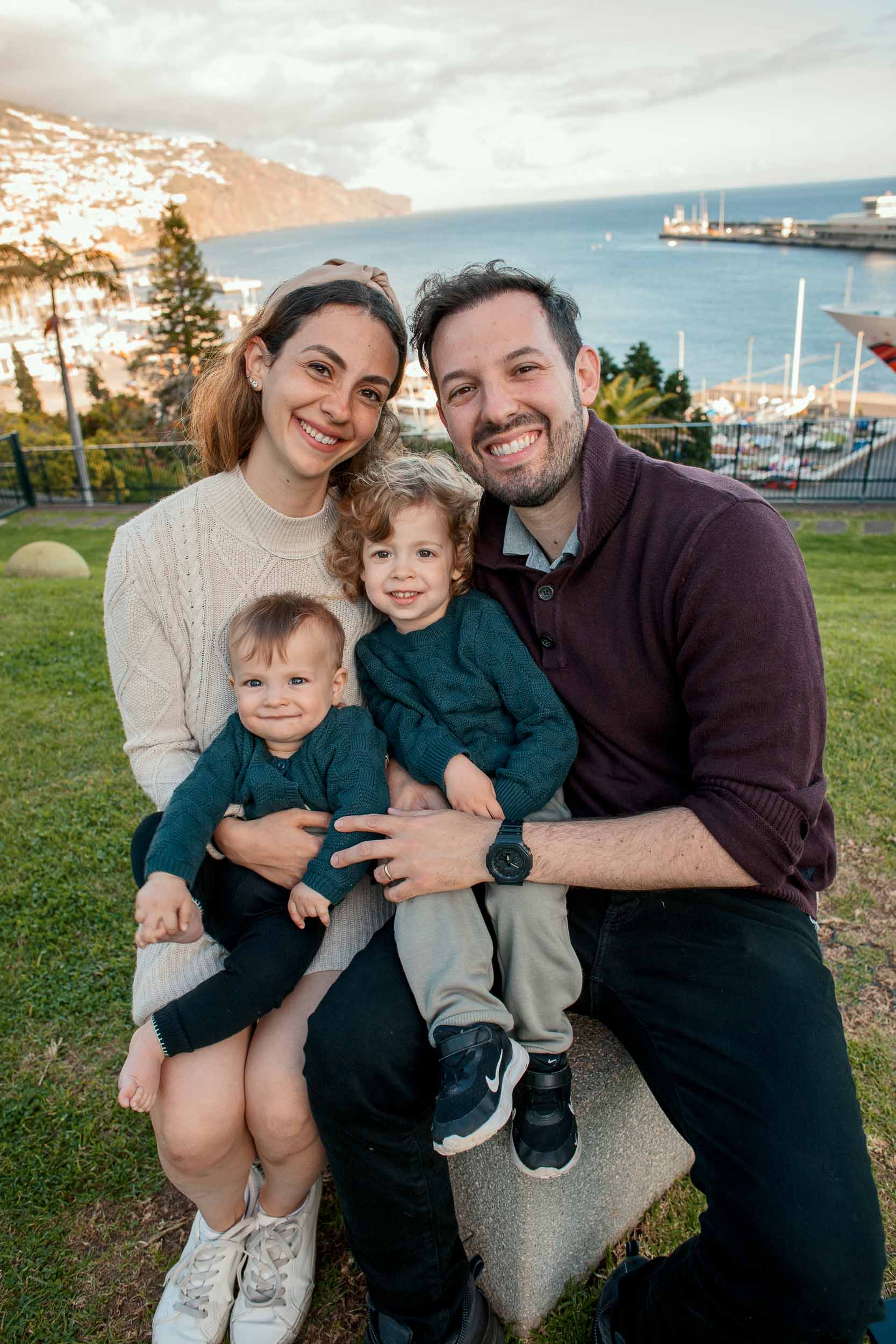 Family of four seated on stone step at Santa Catarina Park, mother holding baby, father holding toddler, Funchal harbor and marina behind, pink evening sky, Madeira