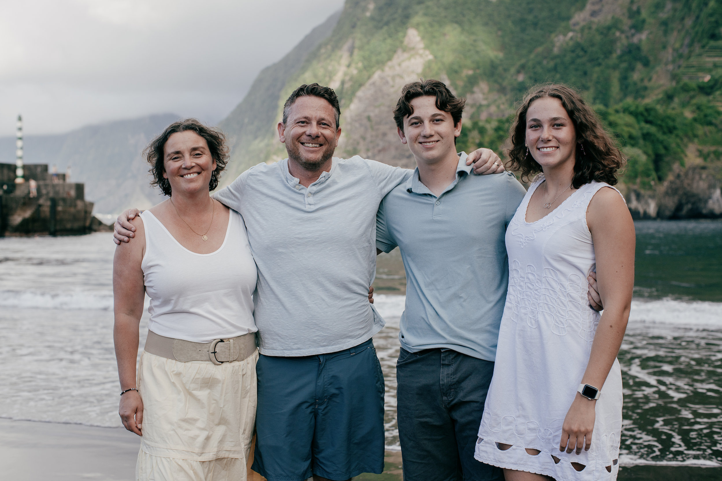 Family of four arm in arm on the black sand at Seixal Beach, green cliffs behind, Madeira