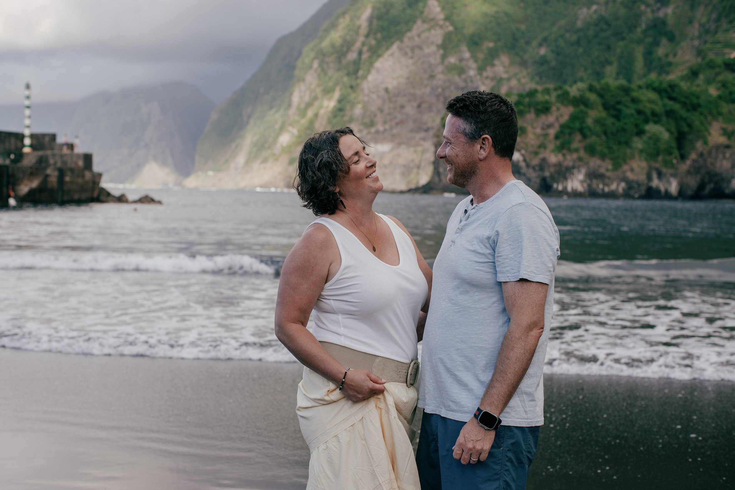 Parents laughing together at the water's edge, harbor pier behind, Seixal, Madeira