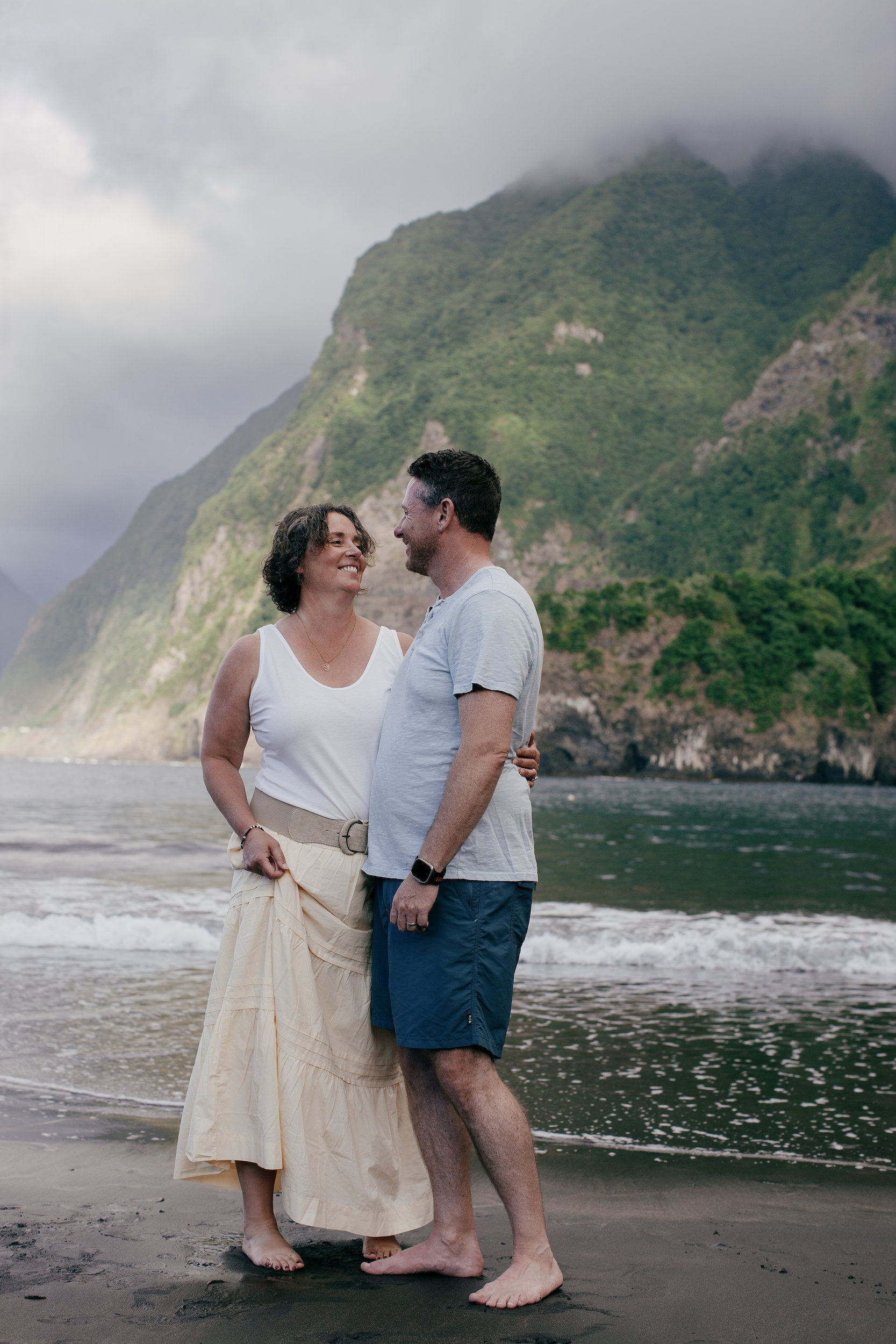 Barefoot on the dark sand together, cliff face green behind, Seixal Beach, Madeira