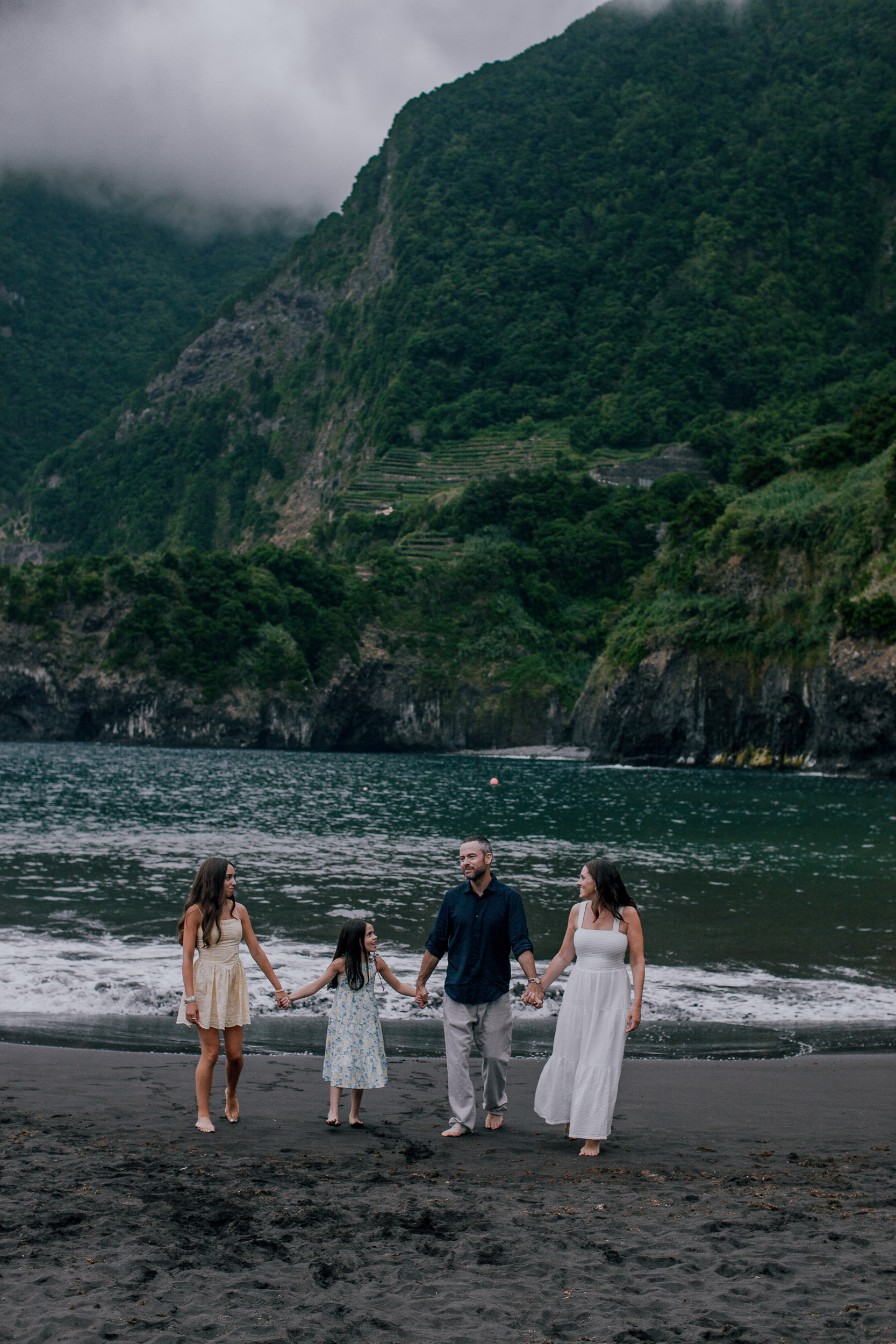 Family of four holding hands at the waterline on an overcast evening, Seixal Beach, Madeira