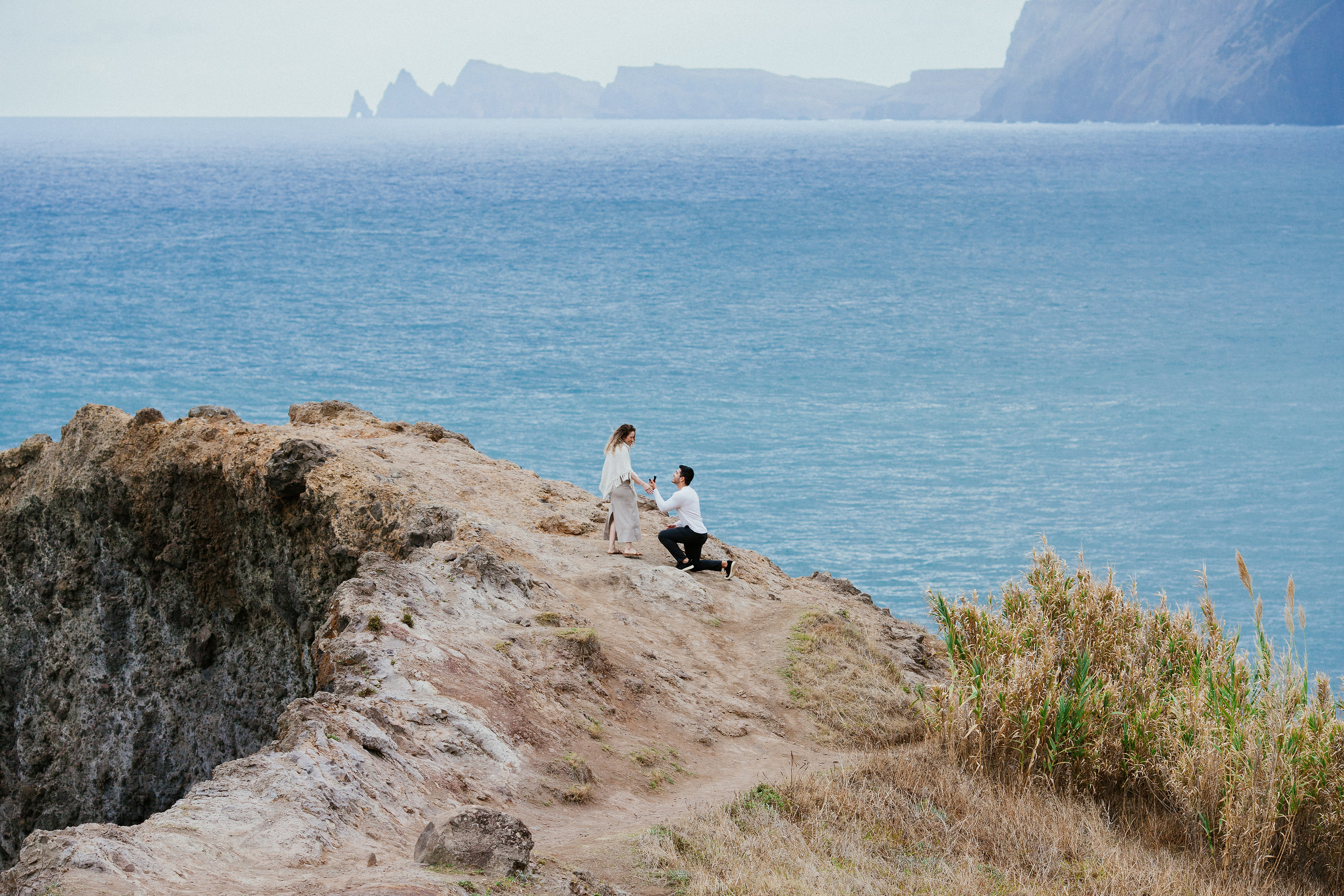 Lucas down on one knee on the rocky ledge, Isabela standing in front of him, the Atlantic behind, Miradouro do Guindaste, Madeira