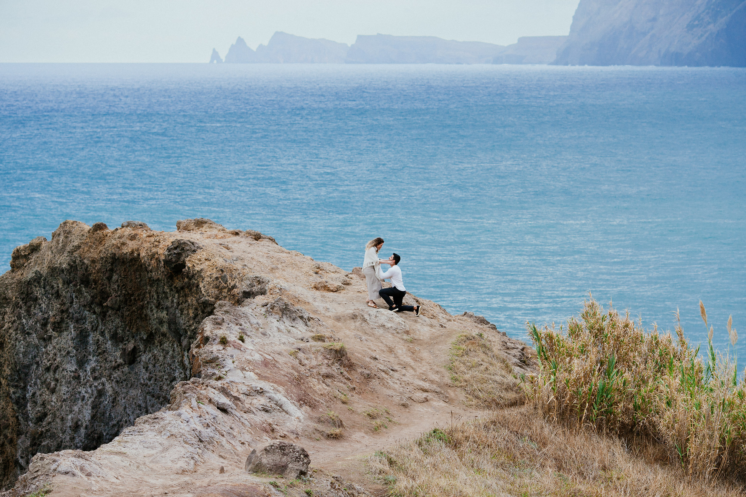 Isabela leaning toward Lucas as he kneels on the cliff ledge, dry grass in the foreground, Miradouro do Guindaste, Madeira