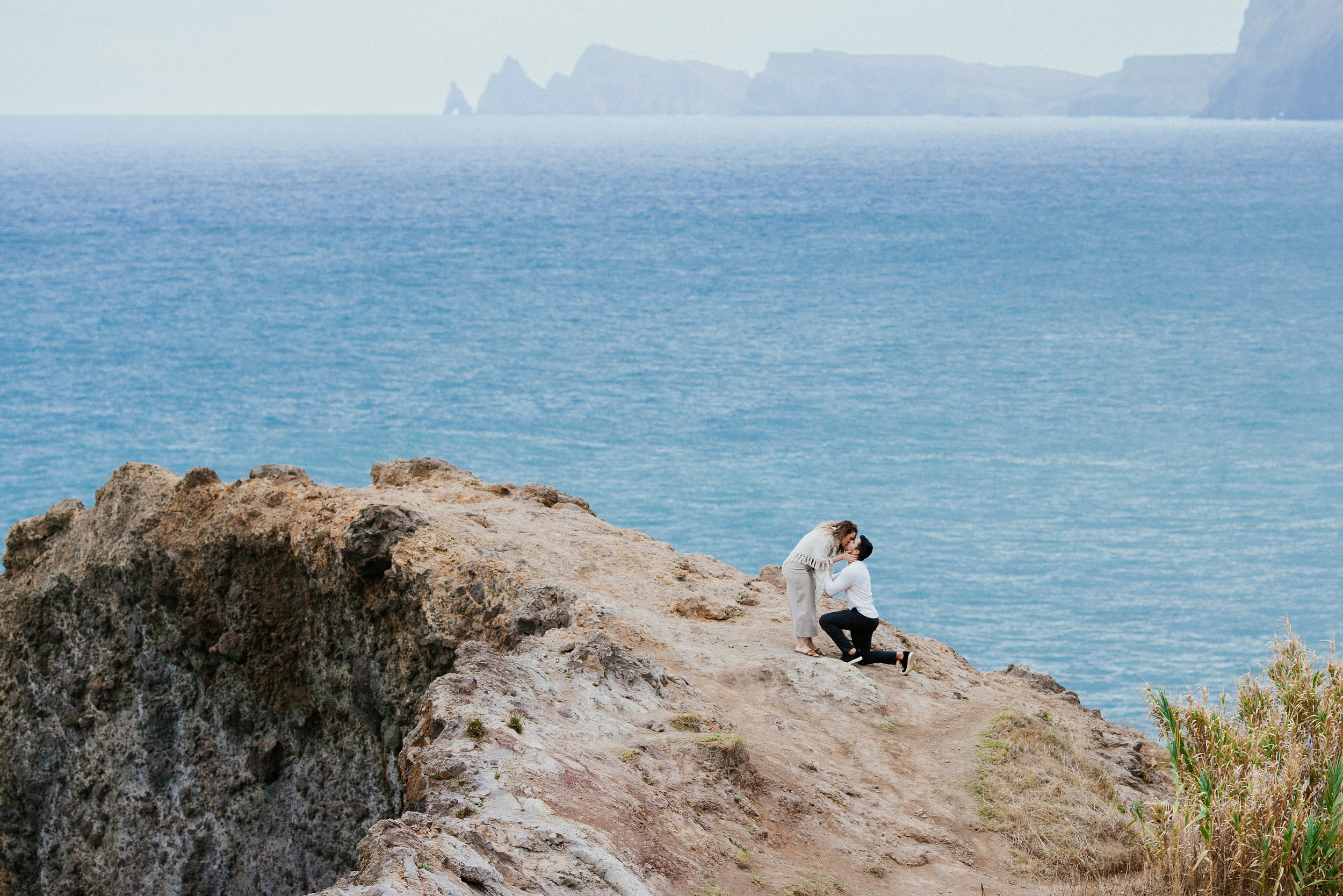 Isabela bending down to kiss Lucas while he is still on one knee, the ocean behind them, Miradouro do Guindaste, Madeira