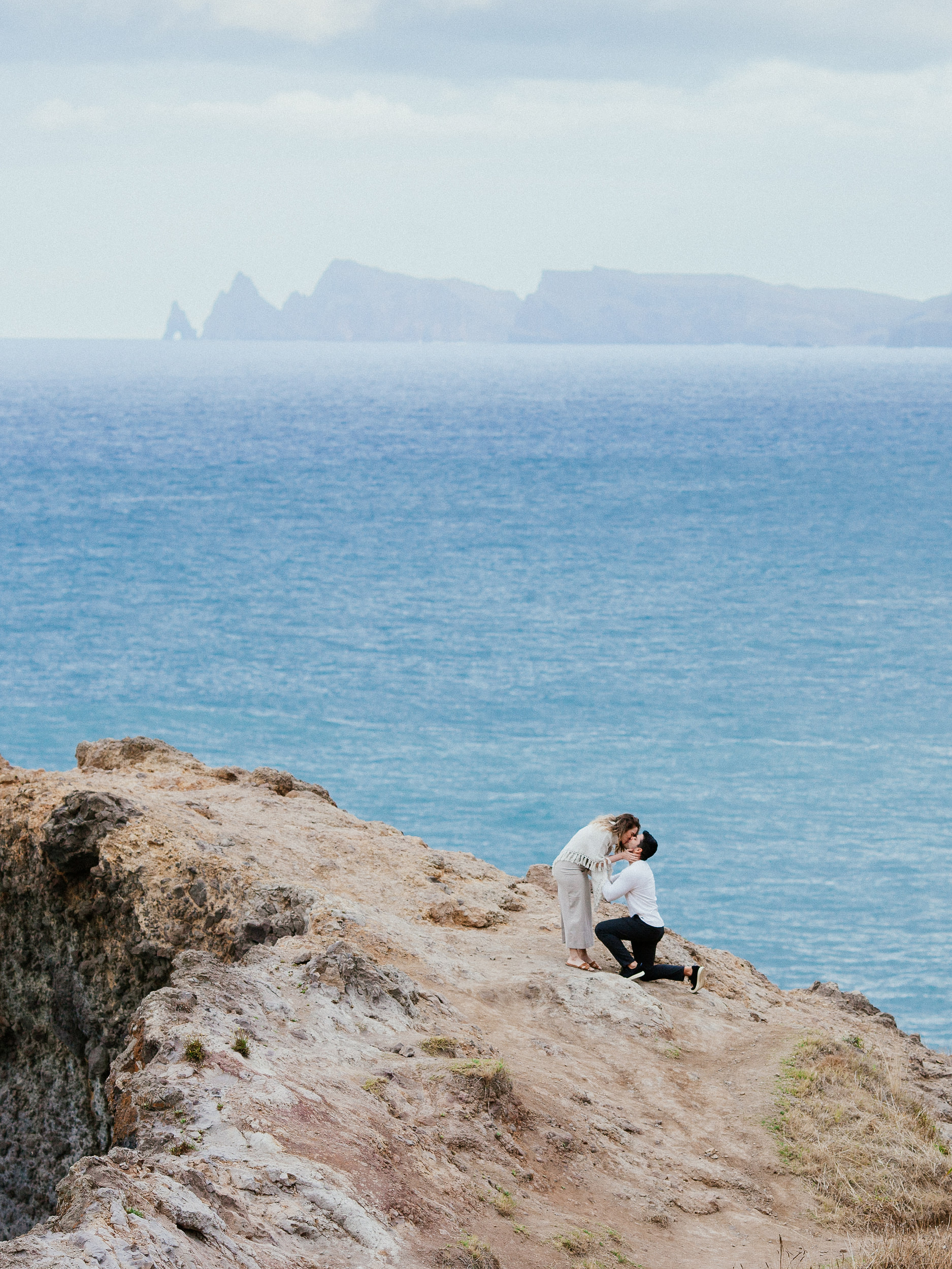 Vertical frame of the kiss on the ledge with Lucas kneeling, overcast sky over the sea, Miradouro do Guindaste, Madeira