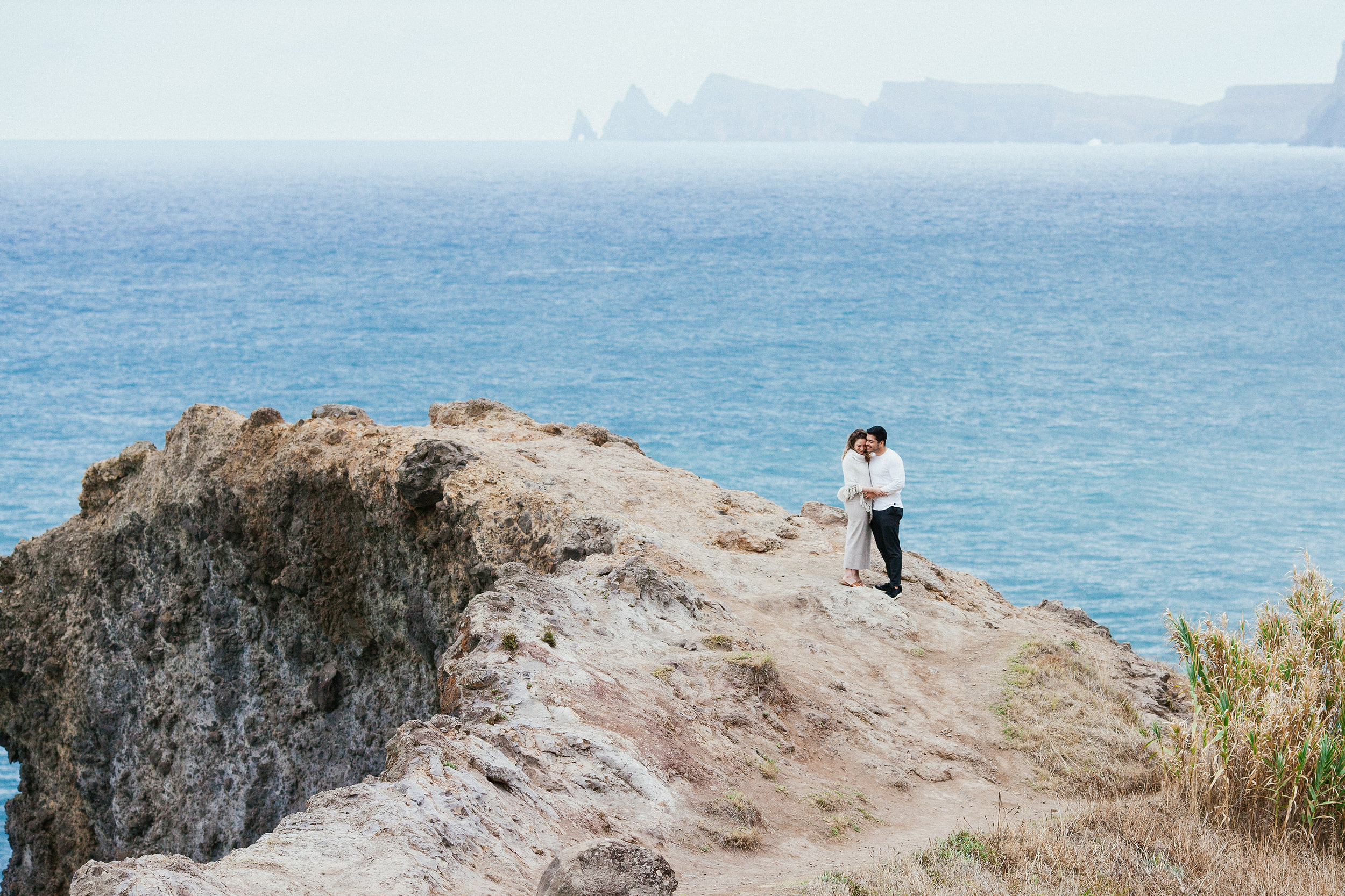 Couple embracing on the cliff ledge, wide view of the Atlantic and far headlands, Miradouro do Guindaste, Madeira