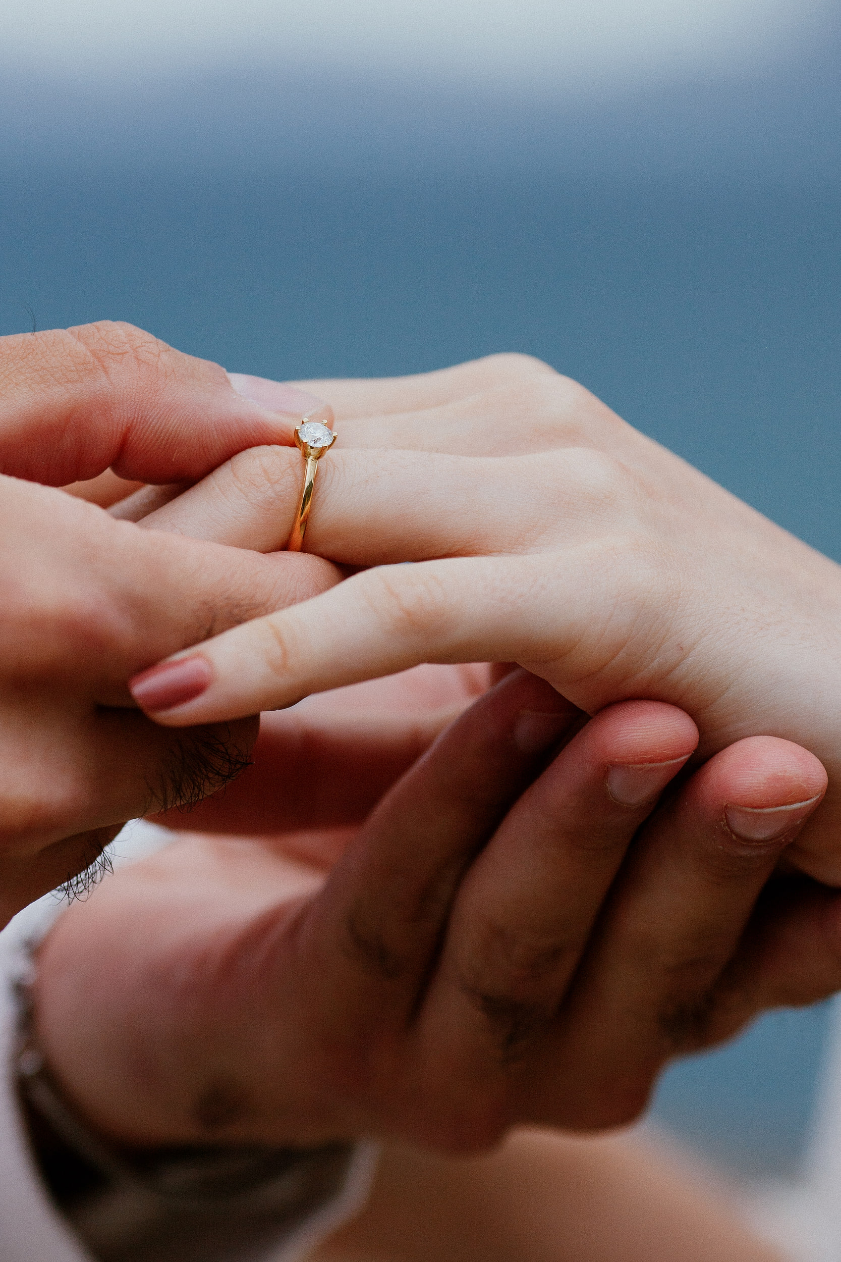 Lucas holding Isabela's fingers, the solitaire ring sharp against the blue sea, Miradouro do Guindaste, Madeira