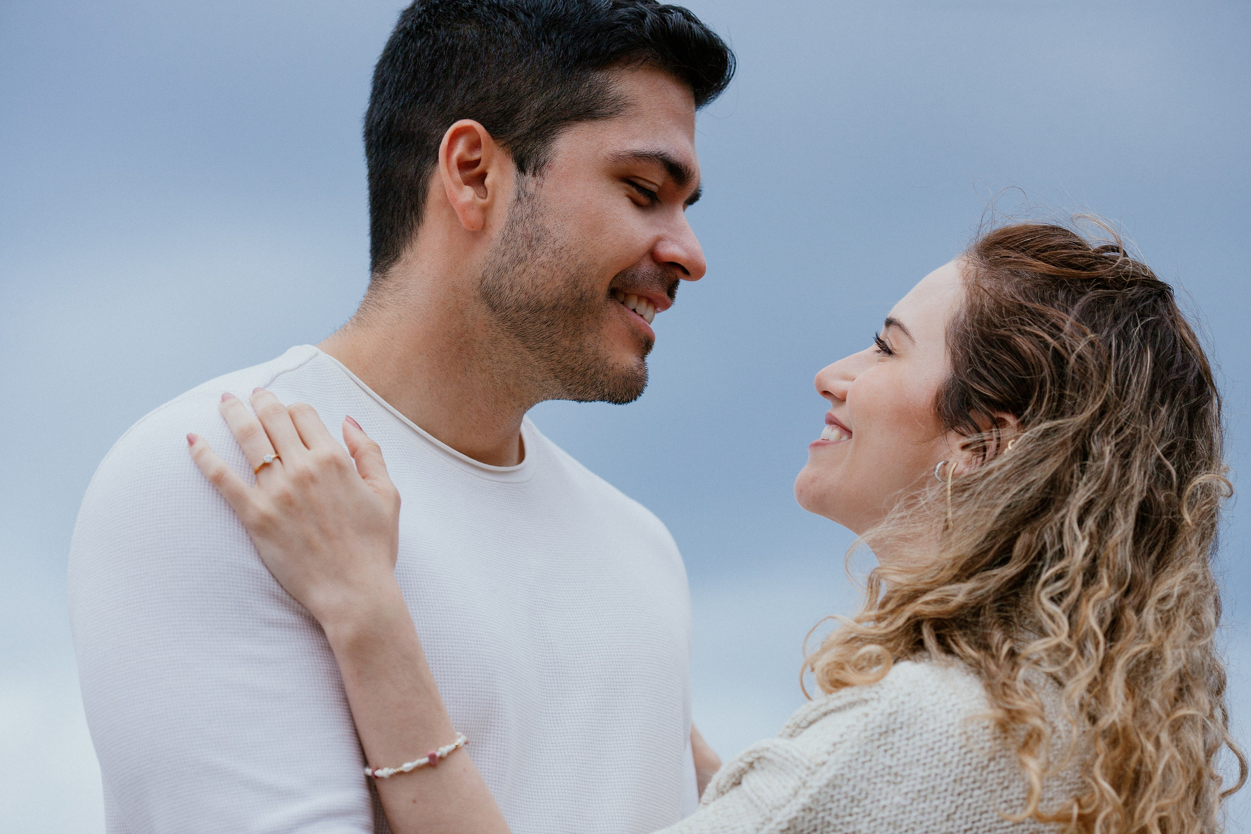 Lucas and Isabela smiling at each other, her ring hand on his shoulder, grey sky behind, Miradouro do Guindaste, Madeira