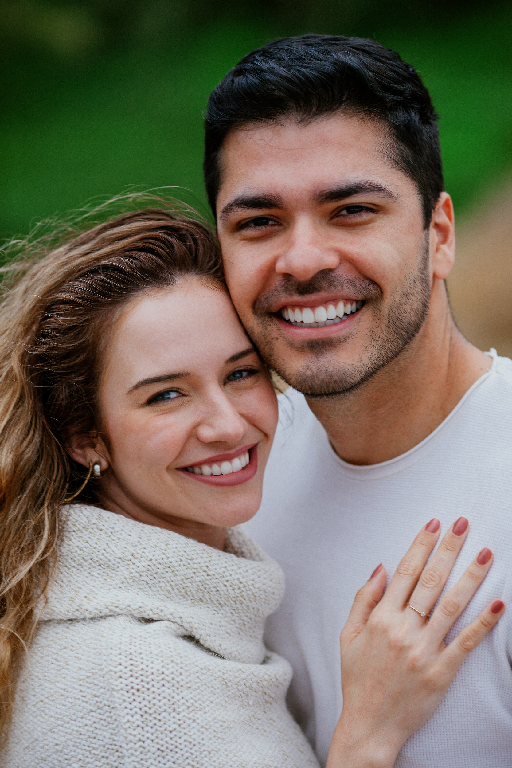 Cheek-to-cheek portrait with Isabela's ring hand on Lucas's chest, green slope blurred behind, Miradouro do Guindaste, Madeira