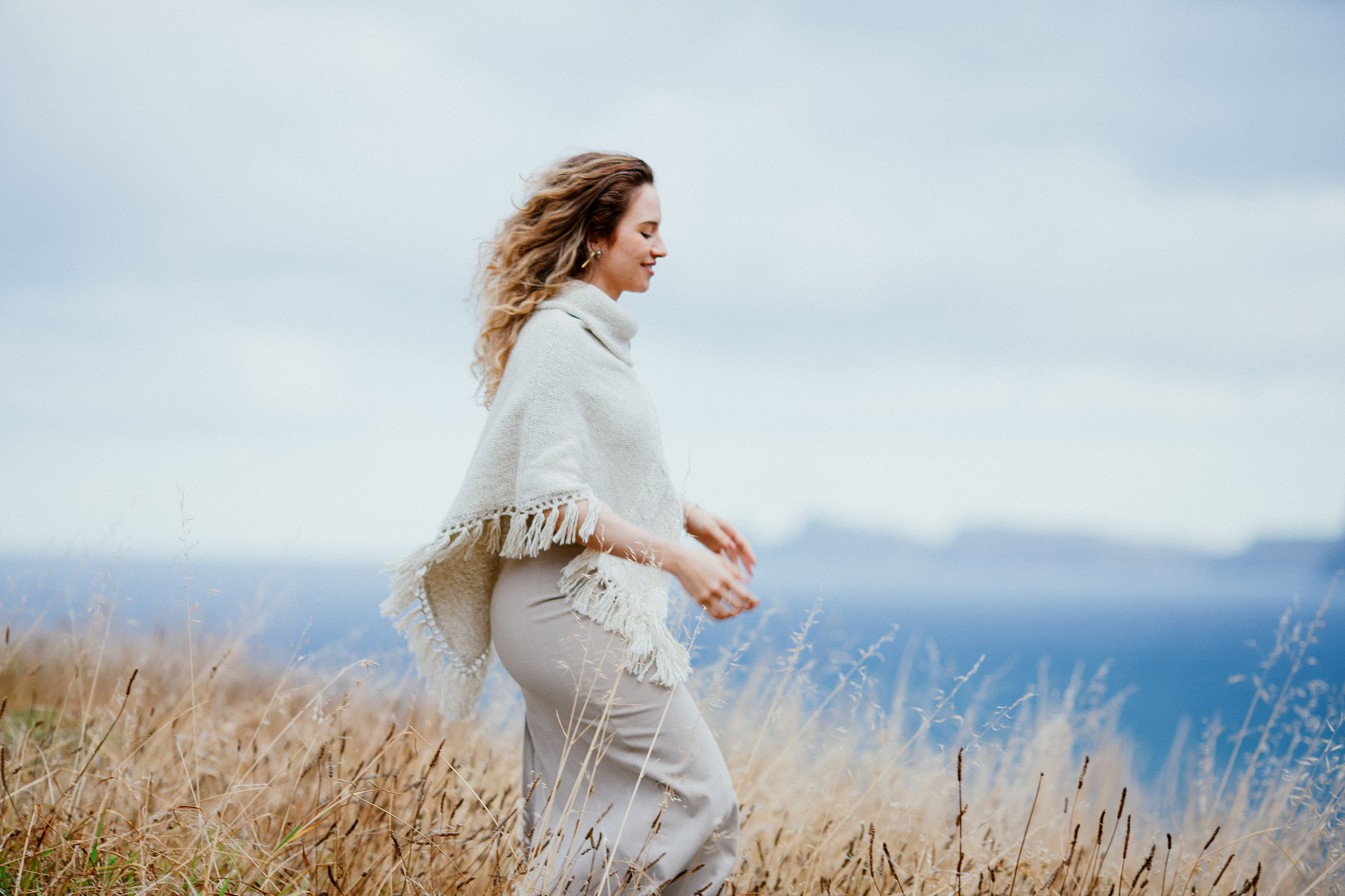 Isabela walking through dry grass above the sea in her fringed cream wrap, Miradouro do Guindaste, Madeira