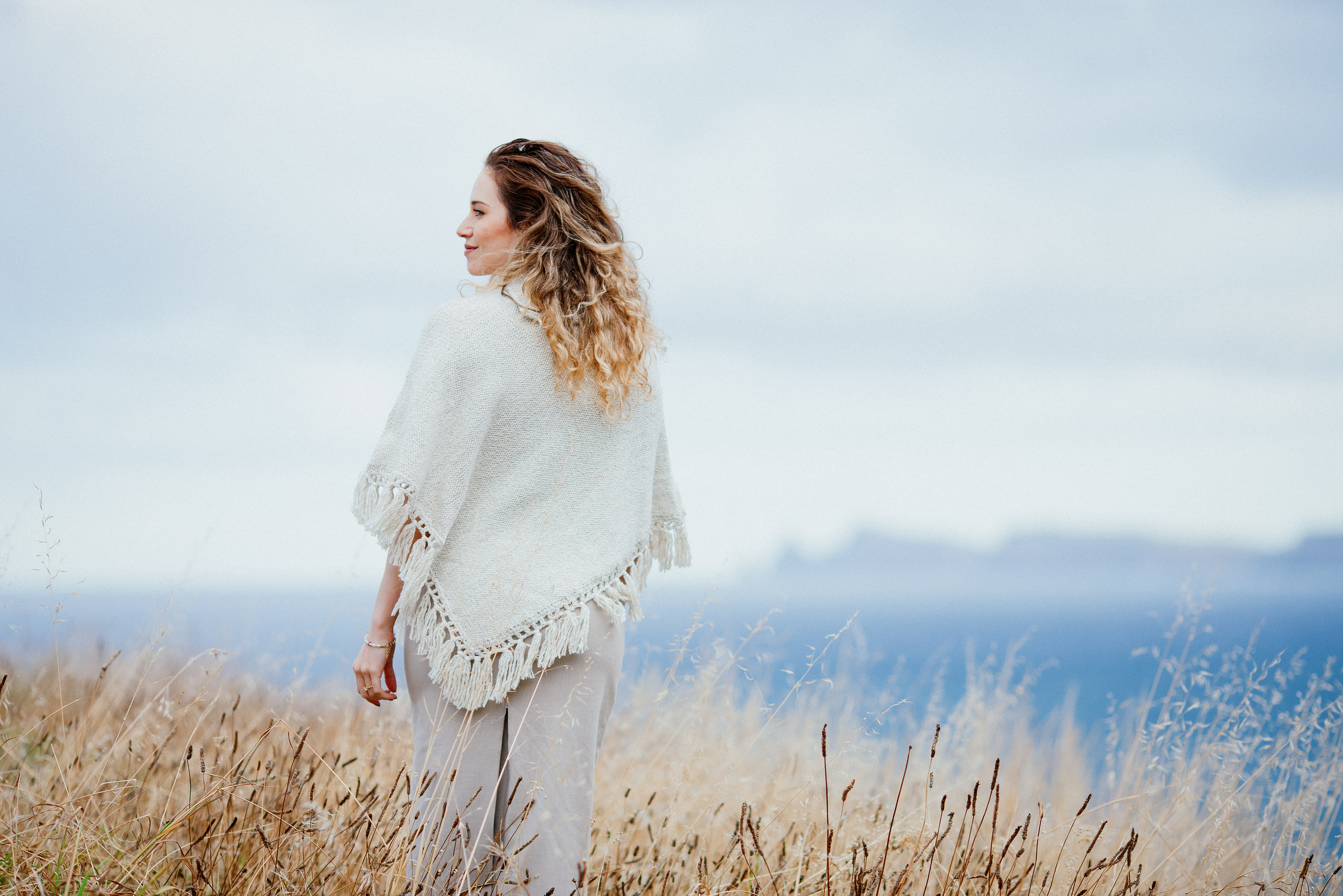 Isabela looking back over her shoulder, dry grass and the ocean behind her, Miradouro do Guindaste, Madeira