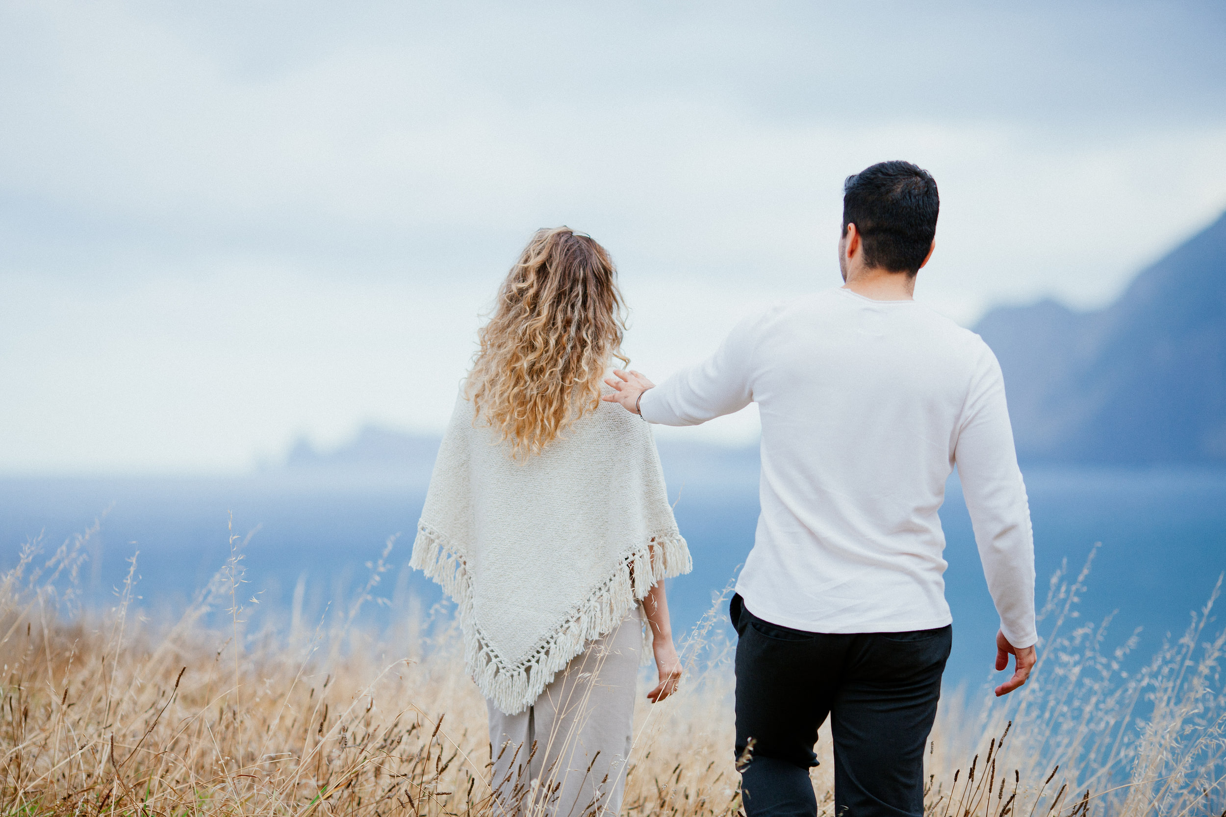 Lucas and Isabela walking away through the grass toward the cliffs, his hand on her shoulder, Miradouro do Guindaste, Madeira