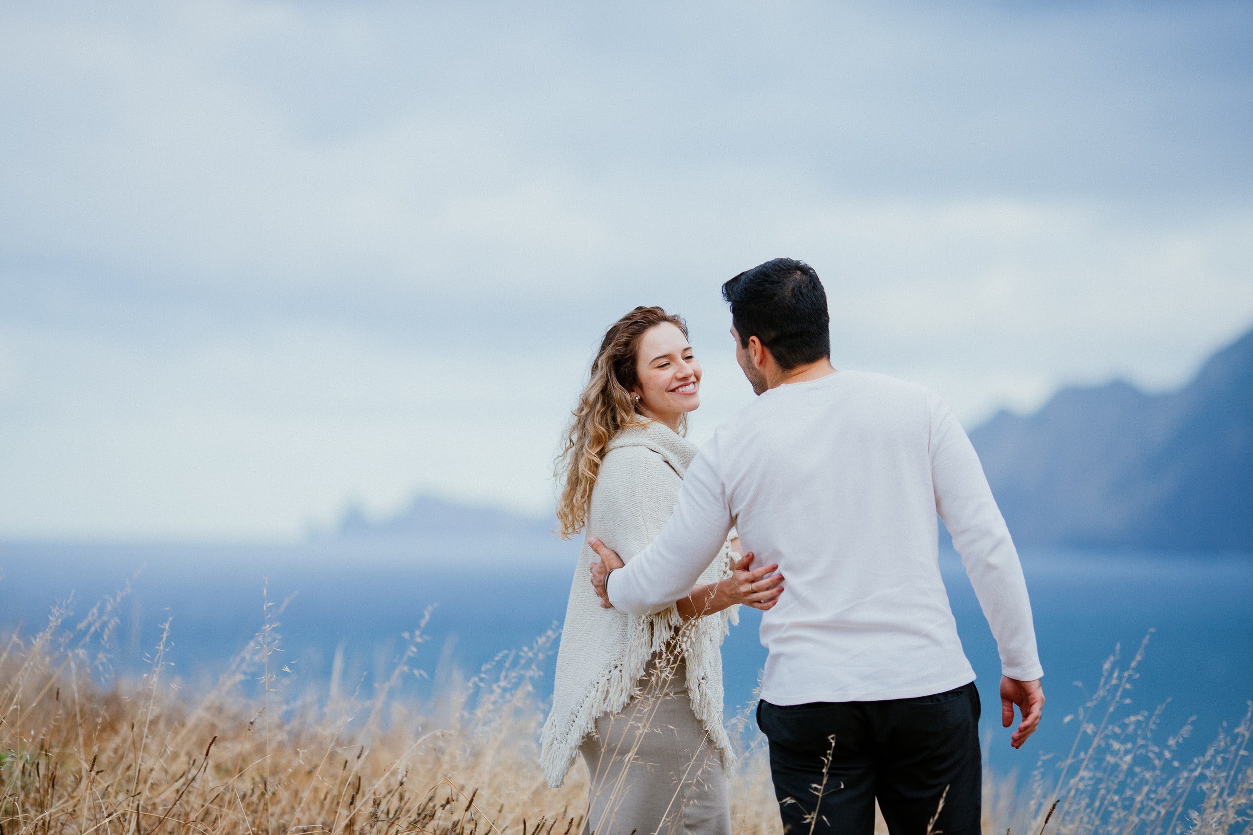 Isabela laughing as Lucas pulls her close while they walk, sea cliffs behind, Miradouro do Guindaste, Madeira