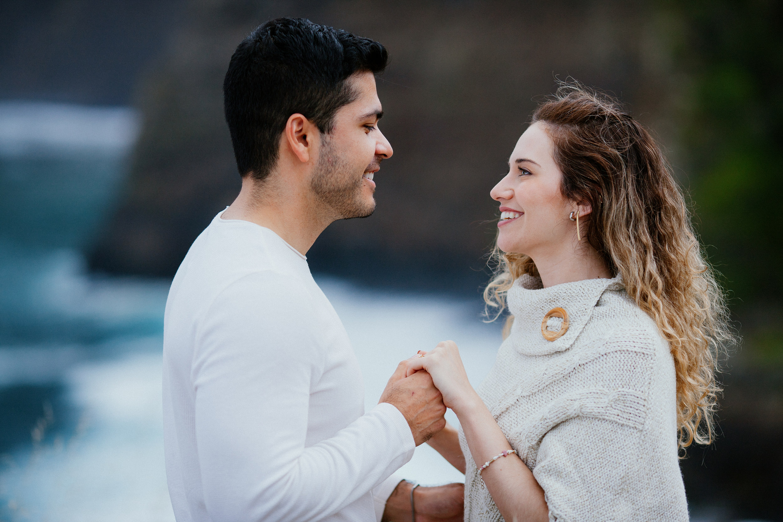 Lucas and Isabela holding hands and smiling at each other, dark cliff and surf behind, Miradouro do Guindaste, Madeira