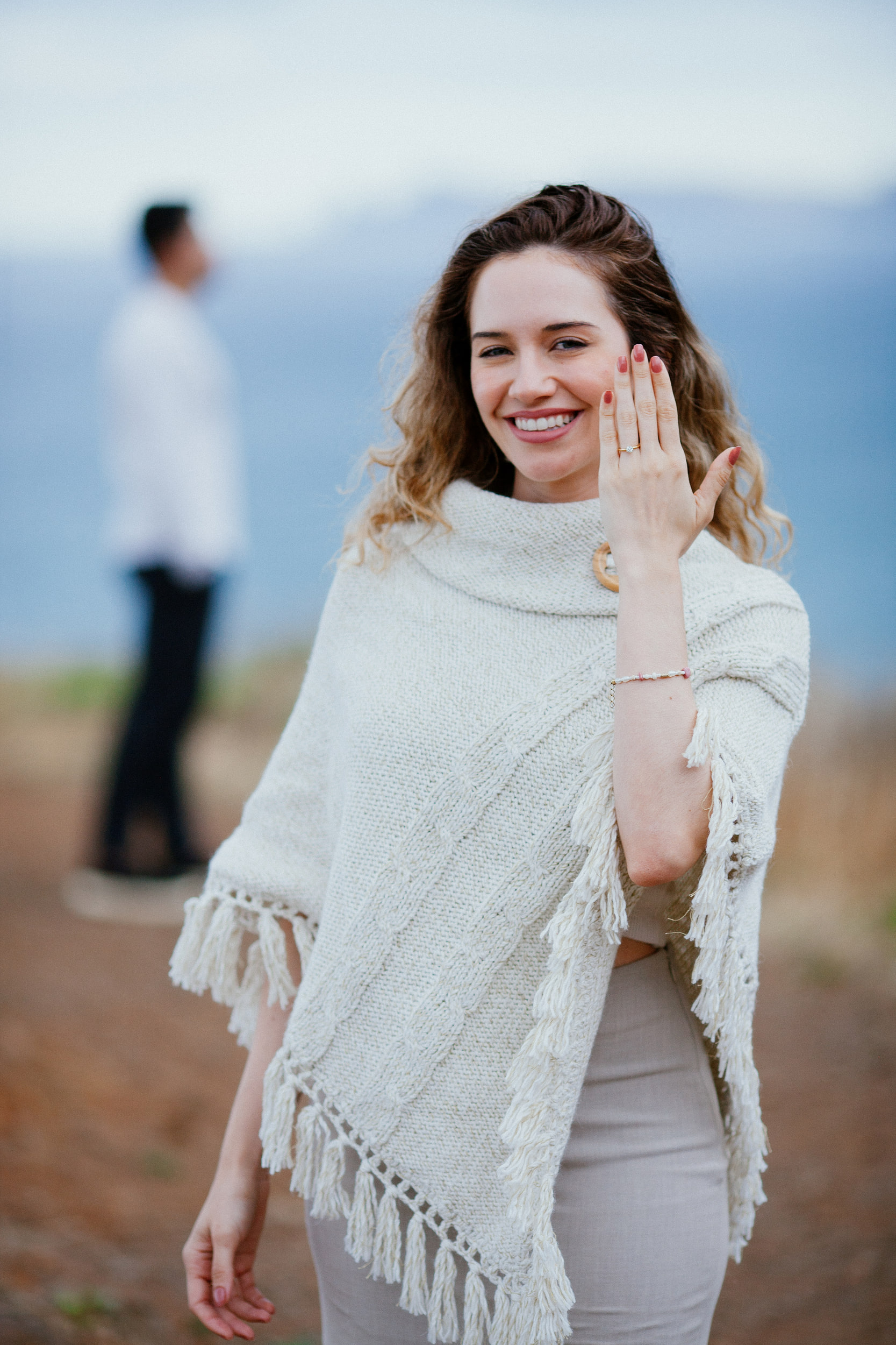 Isabela touching her hair with her ring hand and smiling at the camera, Lucas blurred behind, Miradouro do Guindaste, Madeira