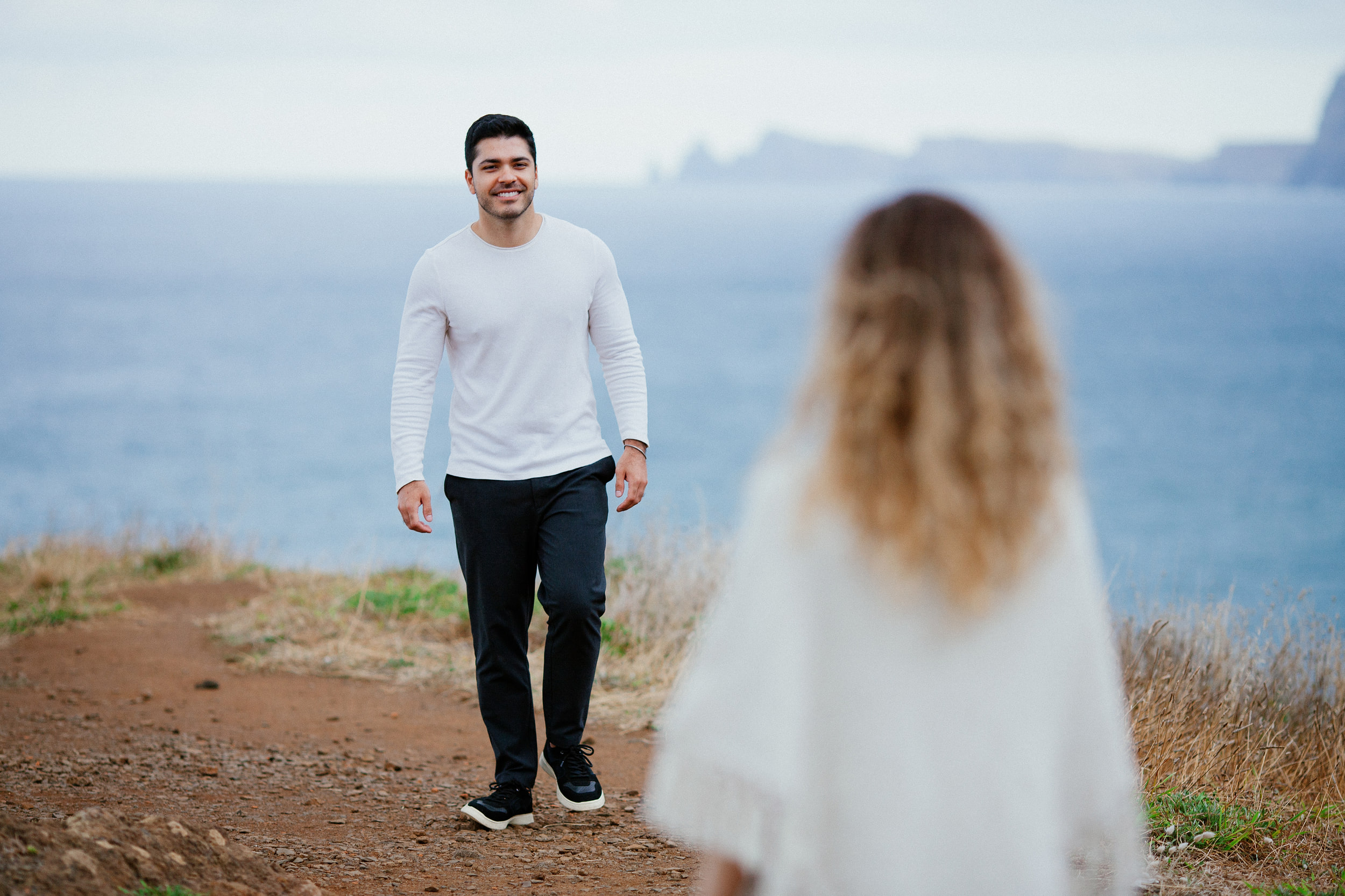 Lucas walking toward Isabela along the dirt path above the sea, smiling, Miradouro do Guindaste, Madeira