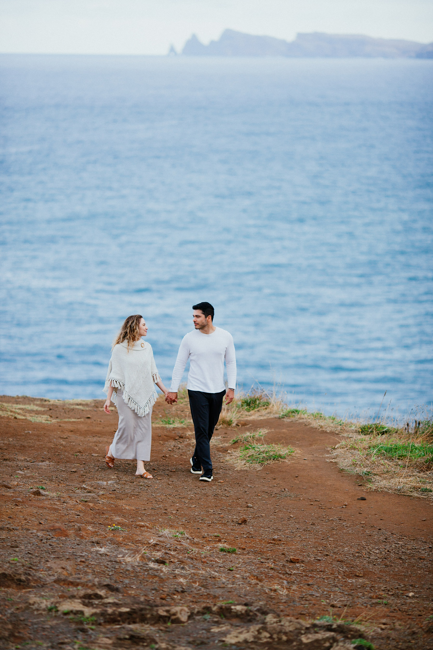 Lucas and Isabela walking hand in hand along the red earth path above the ocean, Miradouro do Guindaste, Madeira