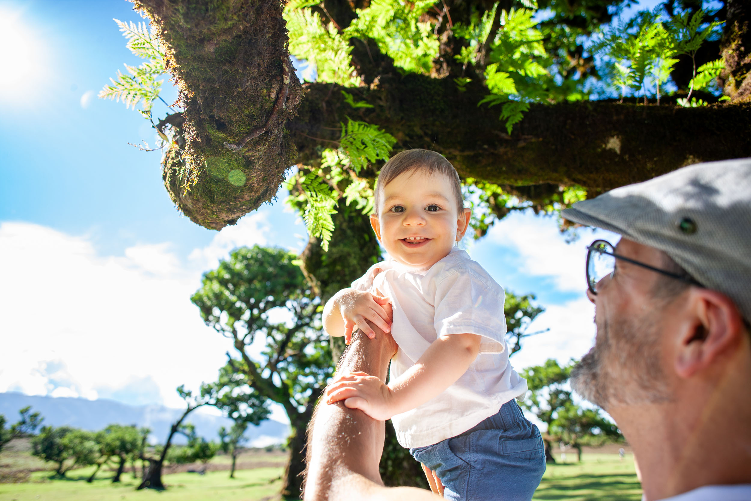 Father holds smiling baby boy beside a moss-covered laurel branch, both looking at each other, blue sky behind, Fanal Forest, Madeira