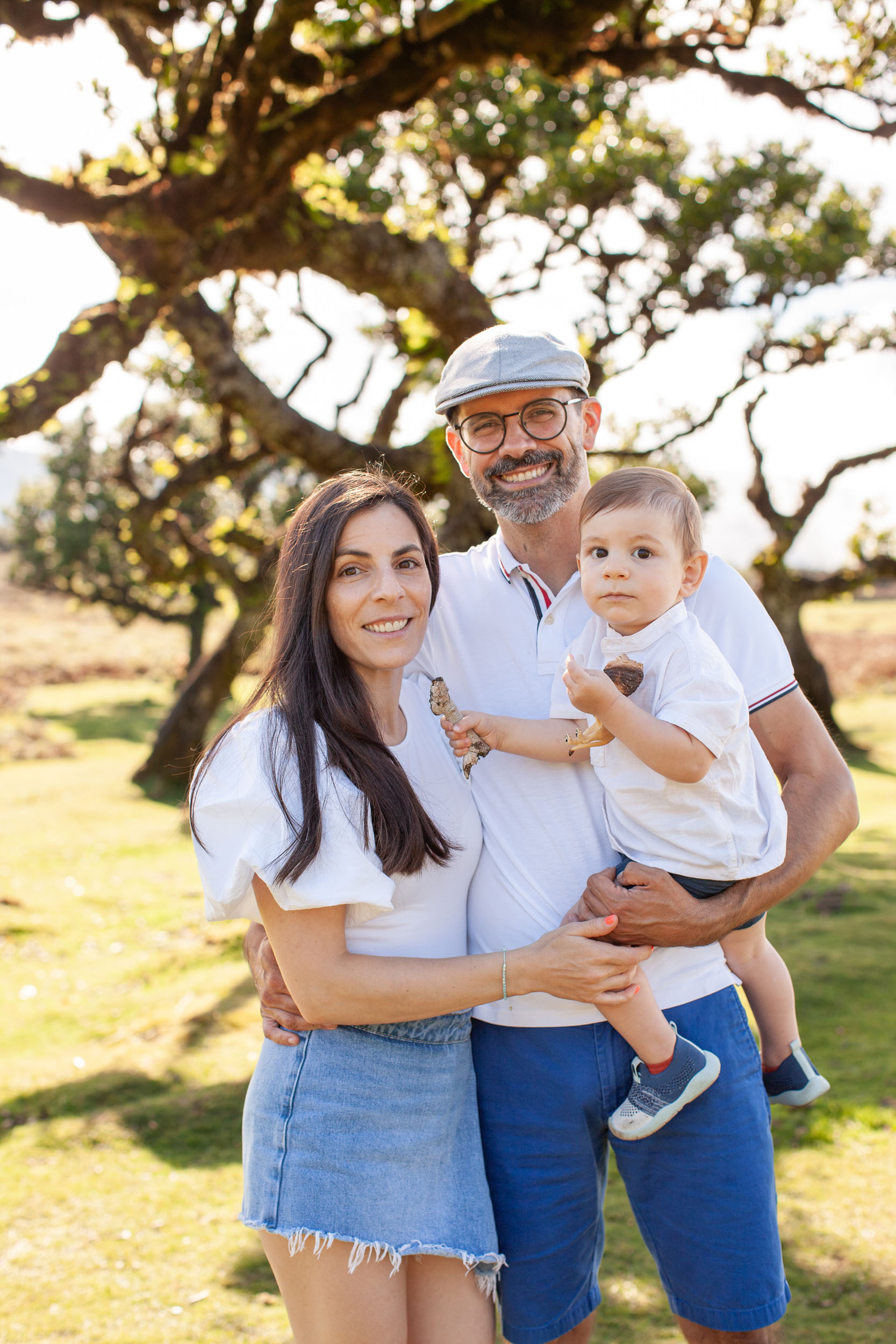 Family portrait in golden afternoon light, mother and father holding baby boy, all smiling at camera, laurel trees behind, Fanal Forest, Madeira