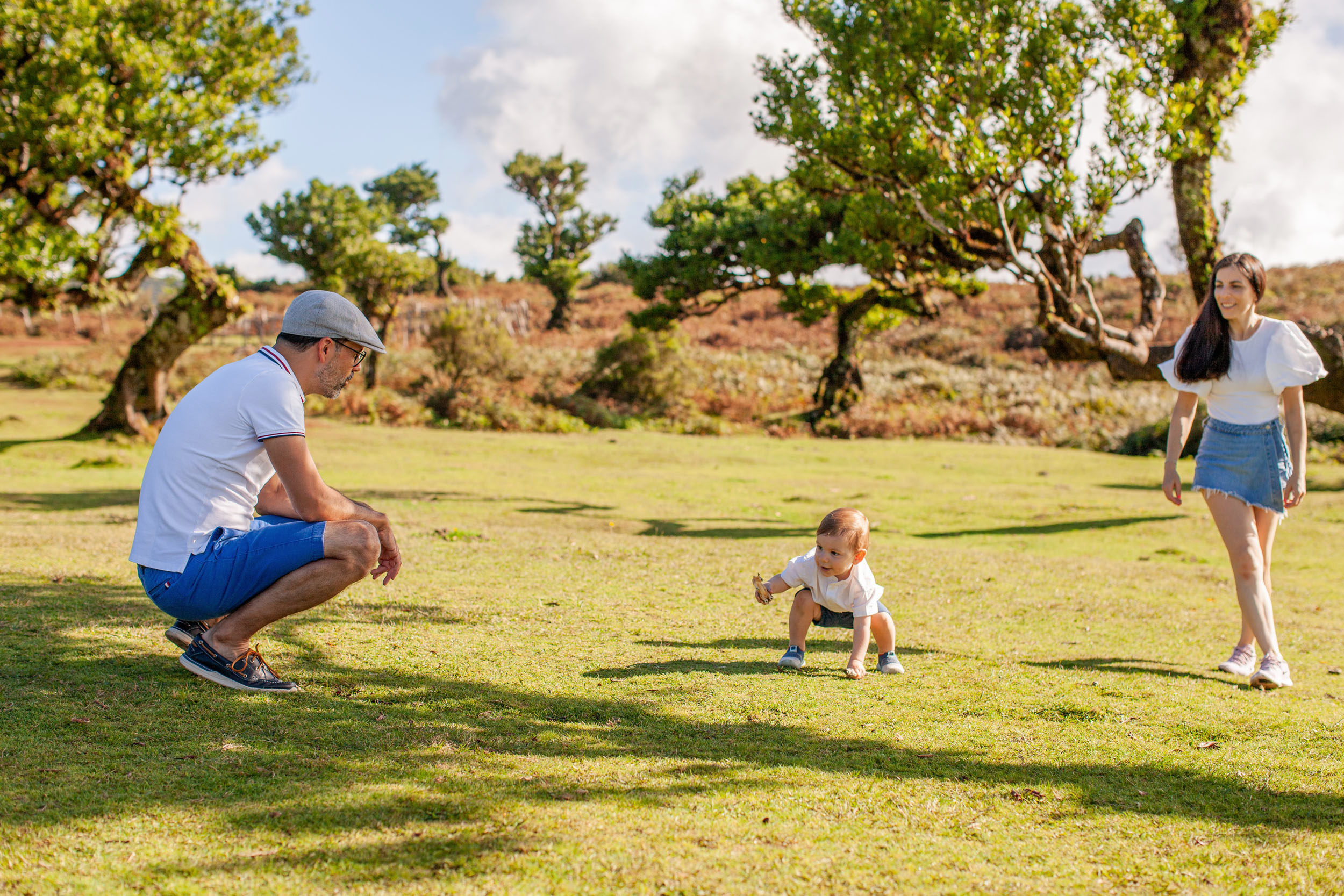 Baby crawls across the green meadow grass, father crouches watching, mother stands smiling, Fanal plateau behind, Madeira